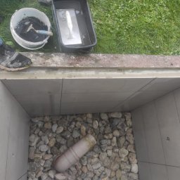 FRW STANISZEWSKI Sp. z.o.o - Wyłożony płytkami dół studzienki kanalizacyjnej z kamiennym dnem, widoczny kubeł z zaprawą, szpachelka i buty robocze na krawędzi.