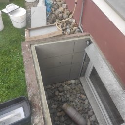 FRW STANISZEWSKI Sp. z.o.o - Wykończone okno piwniczne z szarą okładziną z płytek, drenażem z kamieni i rurą odpływową, widoczne narzędzia i materiały budowlane na zewnątrz.