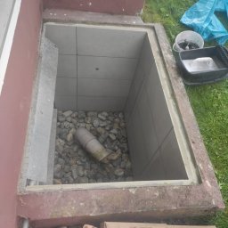 FRW STANISZEWSKI Sp. z.o.o - Otwarty właz do piwnicy wyłożony płytkami z kamienistym dnem i fragmentem rury odpływowej, widoczne narzędzia i materiały budowlane obok.