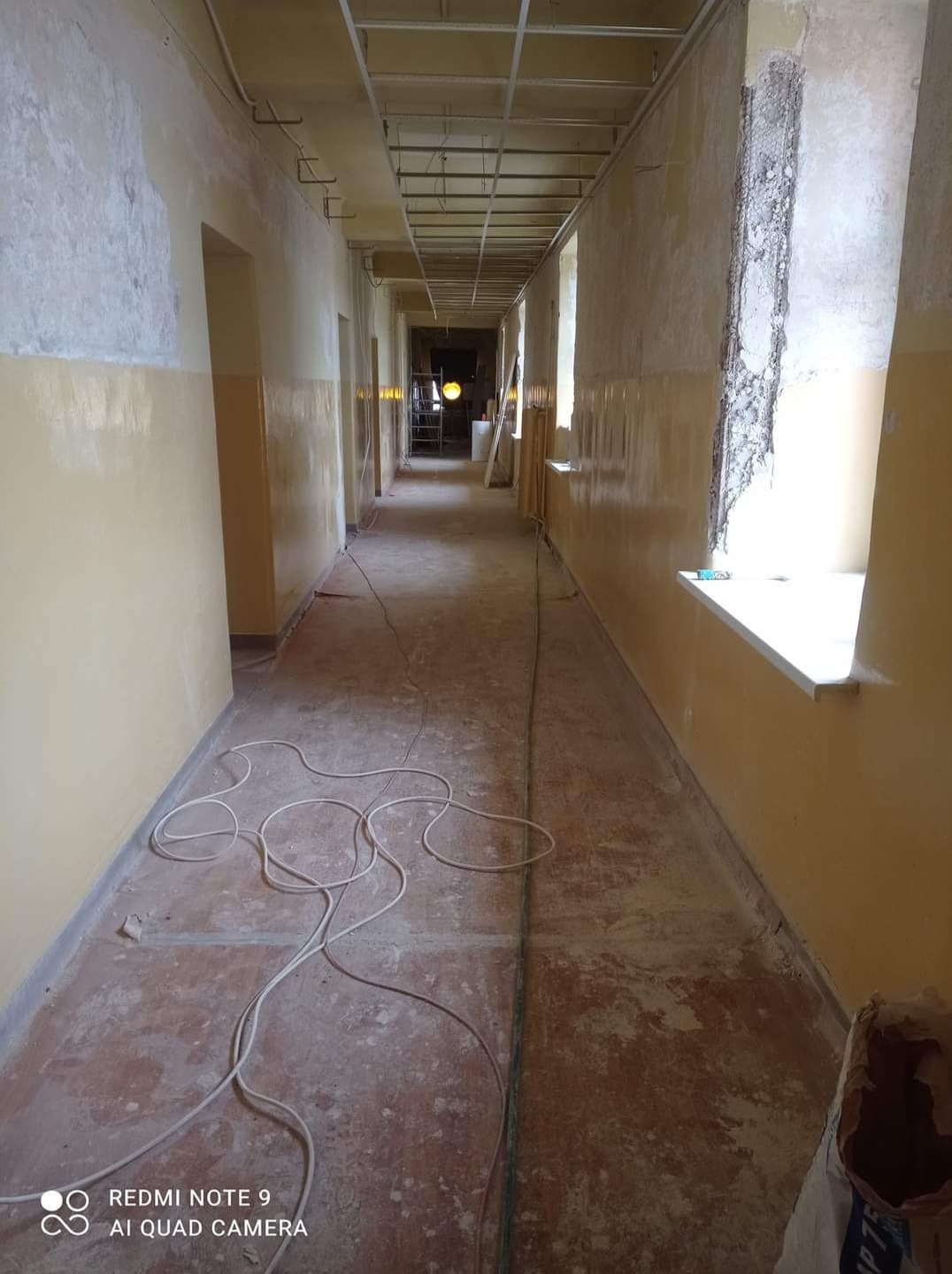 Korytarz w trakcie remontu z widocznymi elementami konstrukcji sufitu podwieszanego, częściowo pomalowanymi ścianami na żółto i wystającymi przewodami elektrycznymi na podłodze.