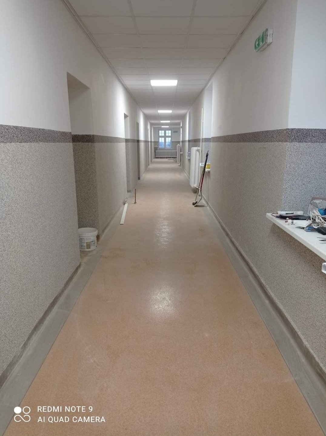 Długi korytarz w budynku biurowym po remoncie: świeżo pomalowane ściany z dwukolorowym wykończeniem, jasna podłoga, widoczne narzędzia malarskie i wiadro z farbą w tle.