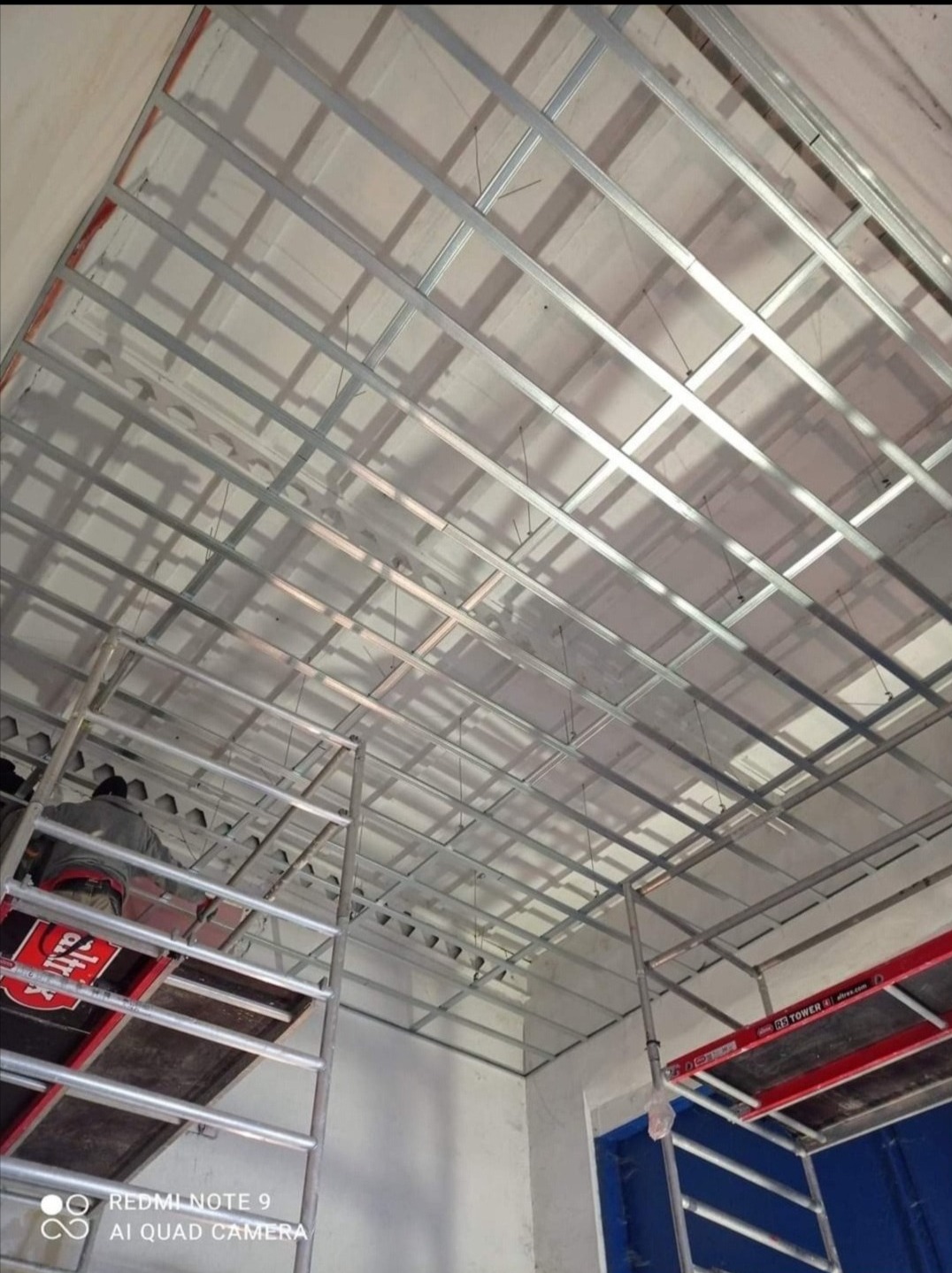 Montaż metalowej konstrukcji sufitu podwieszanego z płyt kartonowo-gipsowych, widok z dołu, fragment rusztowania z czerwoną platformą roboczą po lewej stronie.