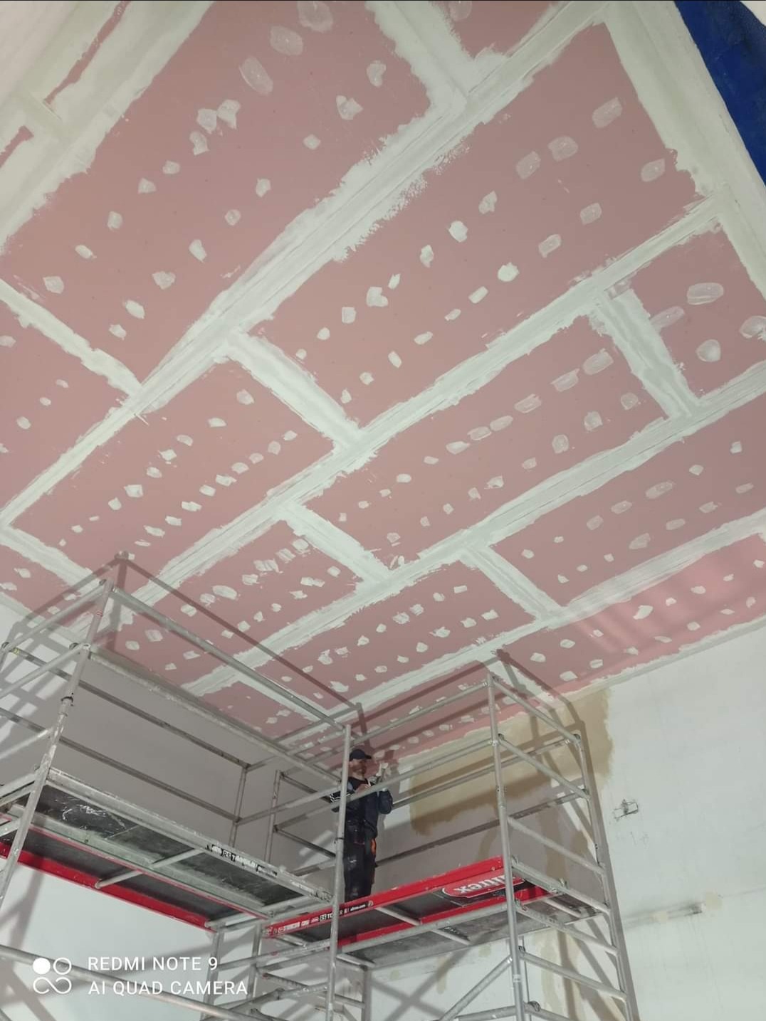 Montaż płyt kartonowo-gipsowych na suficie, widoczne łączenia wypełnione masą szpachlową, pracownik na rusztowaniu.