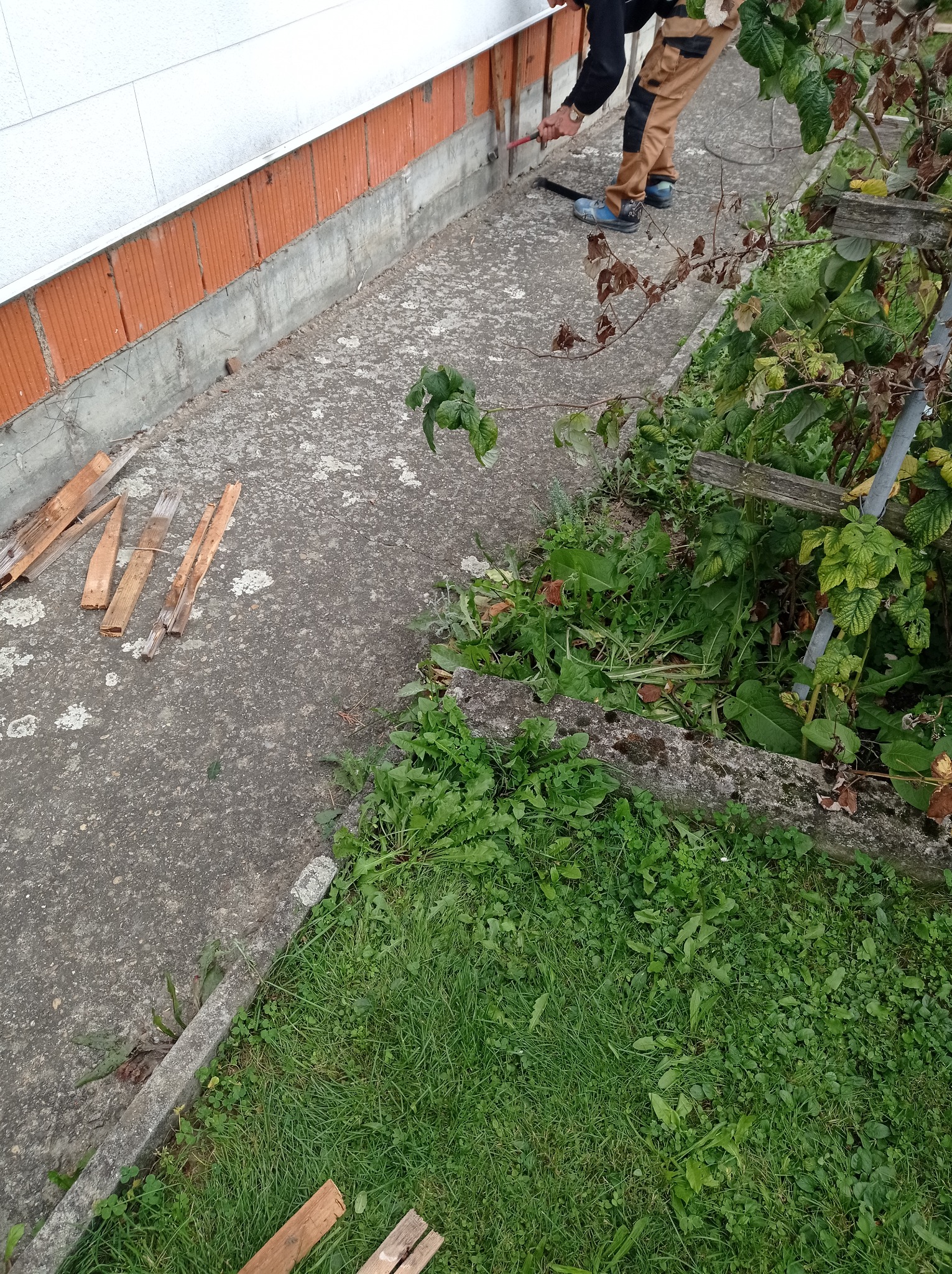 Pracownik w brązowych spodniach i niebieskich butach usuwa resztki zaprawy spod ściany budynku z wykorzystaniem szpachelki i młotka, widoczne fragmenty fundamentów i roślinność.