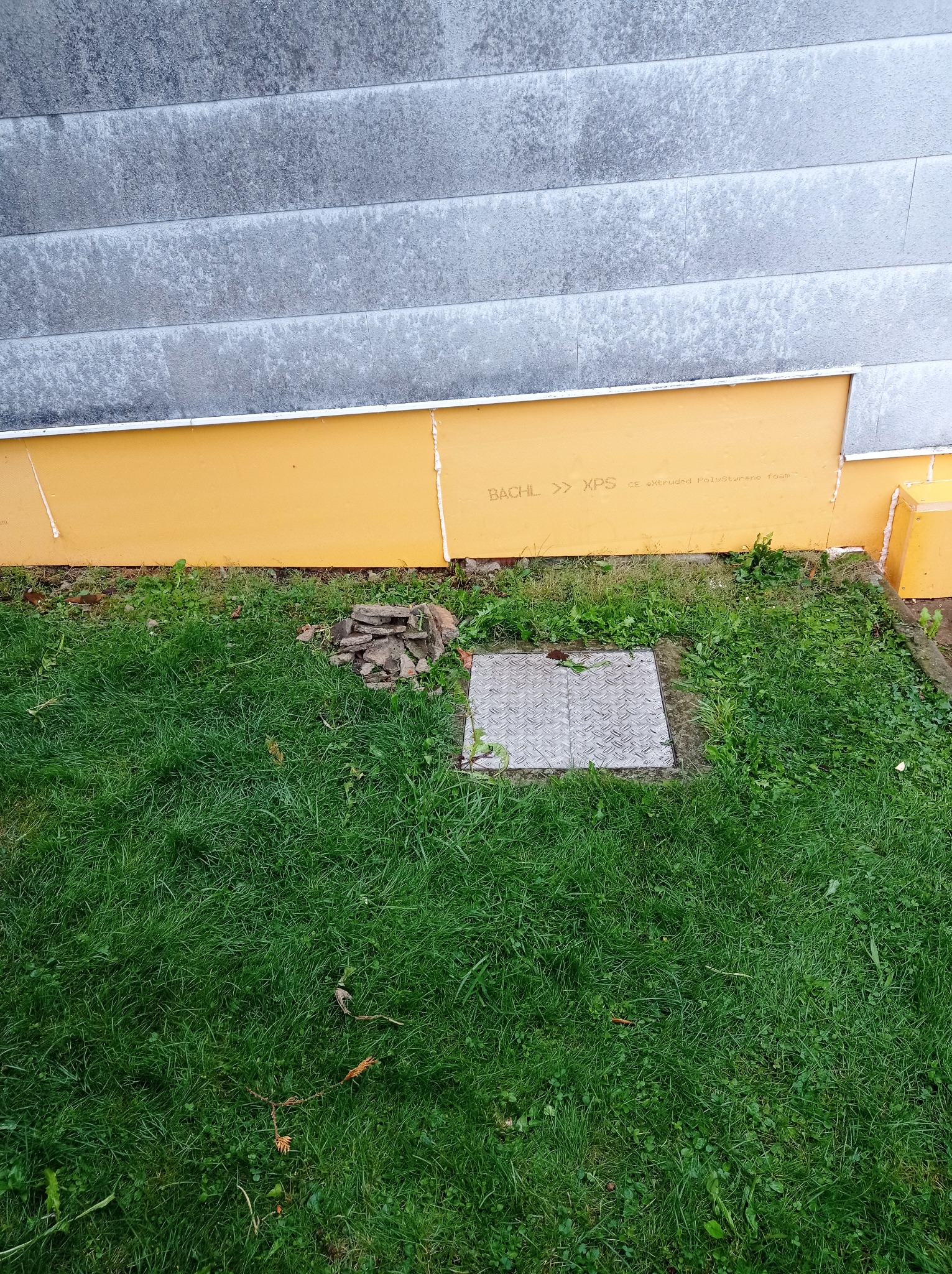 Ocieplenie fundamentu budynku z widoczną żółtą płytą XPS, fragmentem szarej elewacji i trawnikiem z metalową klapą rewizyjną.