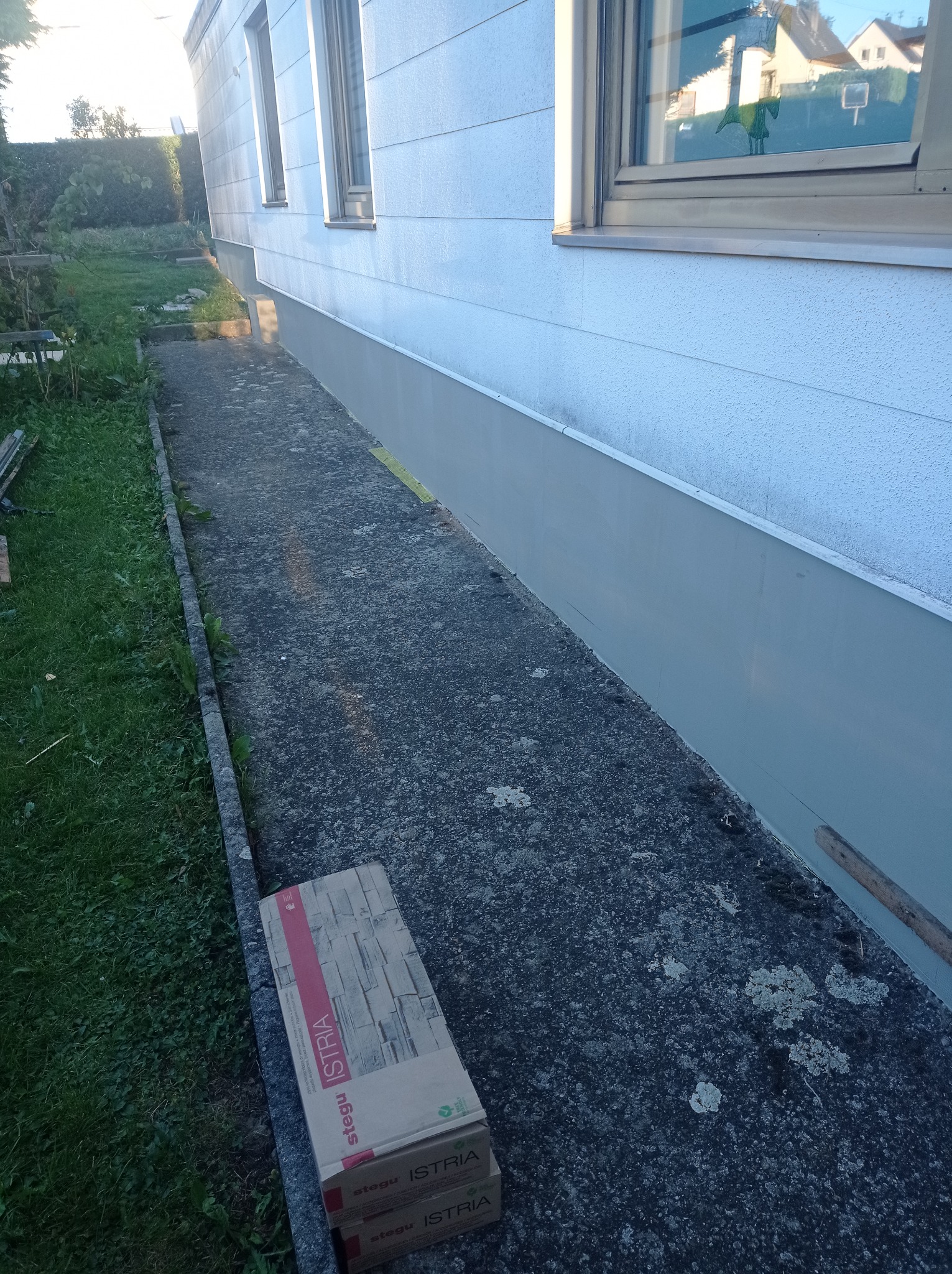 Wykończona elewacja budynku z widocznym cokołem pomalowanym na szaro, obok ułożone kartony z okładziną elewacyjną Stegu Istria.