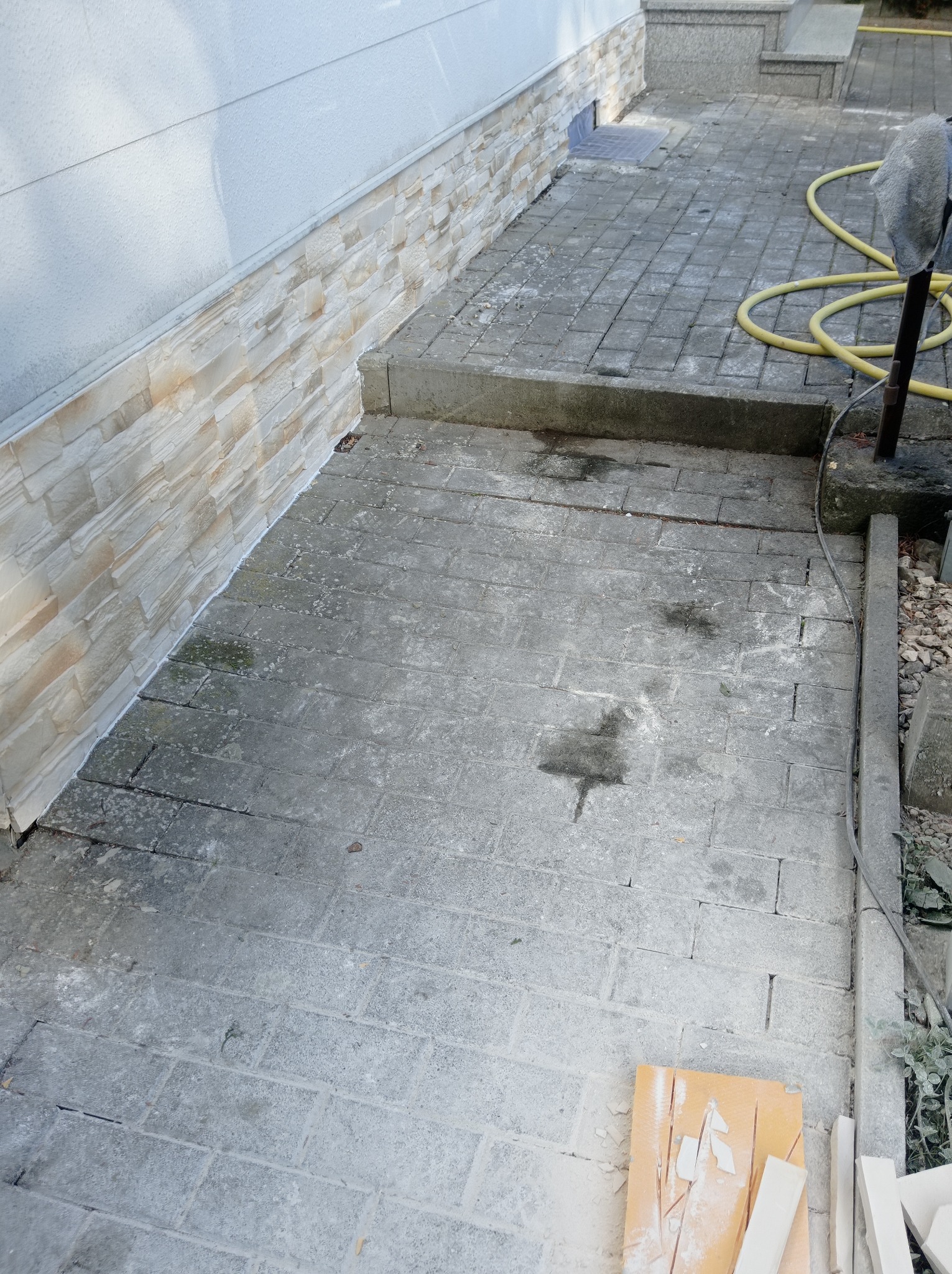 Wykończona elewacja z kamienia dekoracyjnego przy ziemi, z widocznym fragmentem kostki brukowej i żółtym wężem ogrodowym w tle, oraz resztkami materiałów budowlanych leżących na ziemi.