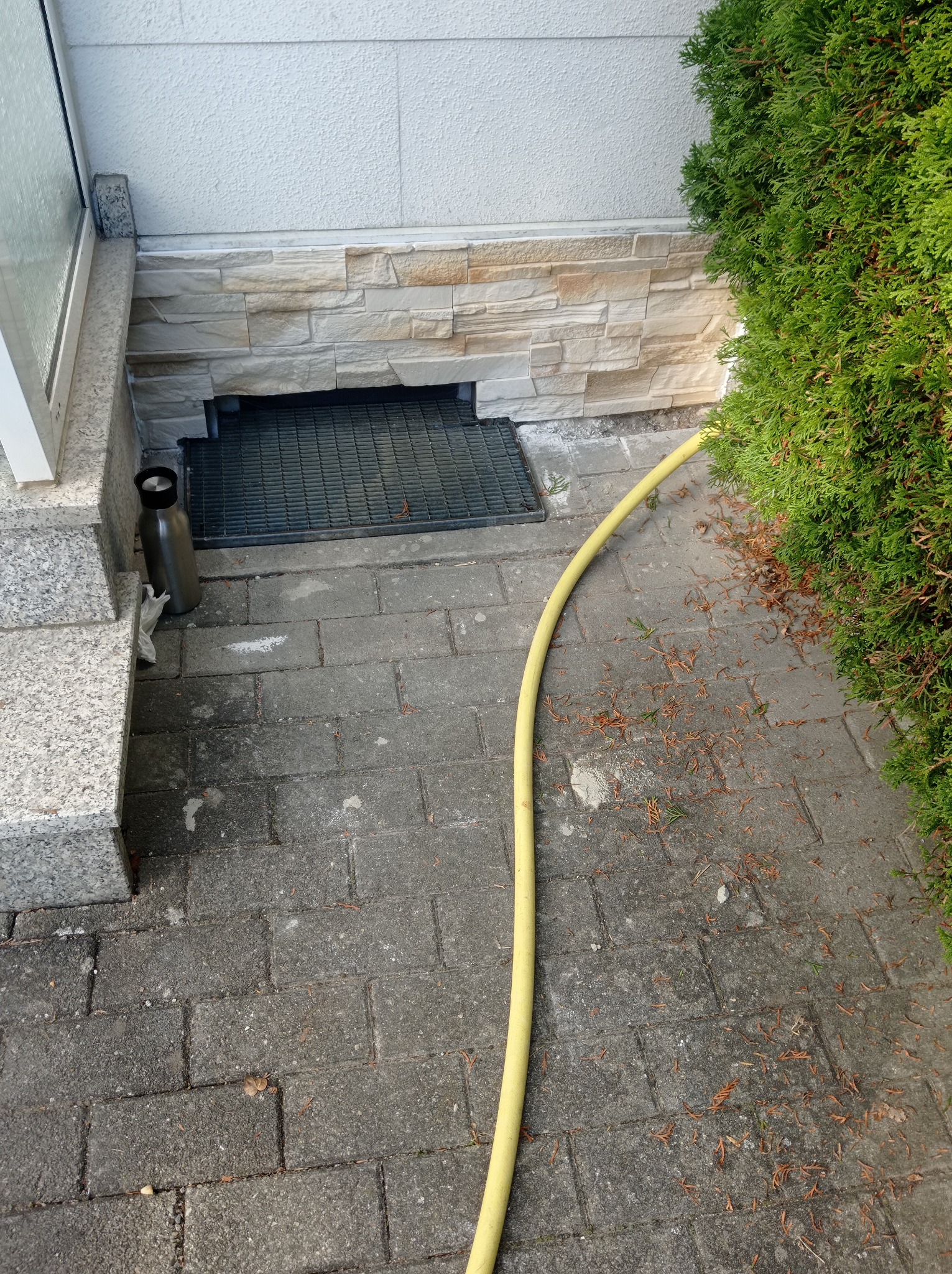 Wykończenie elewacji przy wejściu do budynku z dekoracyjnym kamieniem, kratką wentylacyjną i żółtym wężem ogrodowym na brukowanym chodniku.