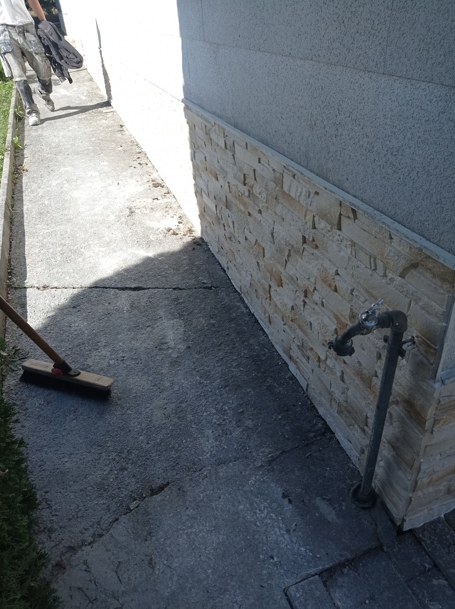 Fragment elewacji budynku wykończony dekoracyjną okładziną z kamienia naturalnego, widoczna instalacja wodna i zamiataczka z drewnianym trzonkiem, w tle pracownik w ubrudzonych spodniach roboczych.