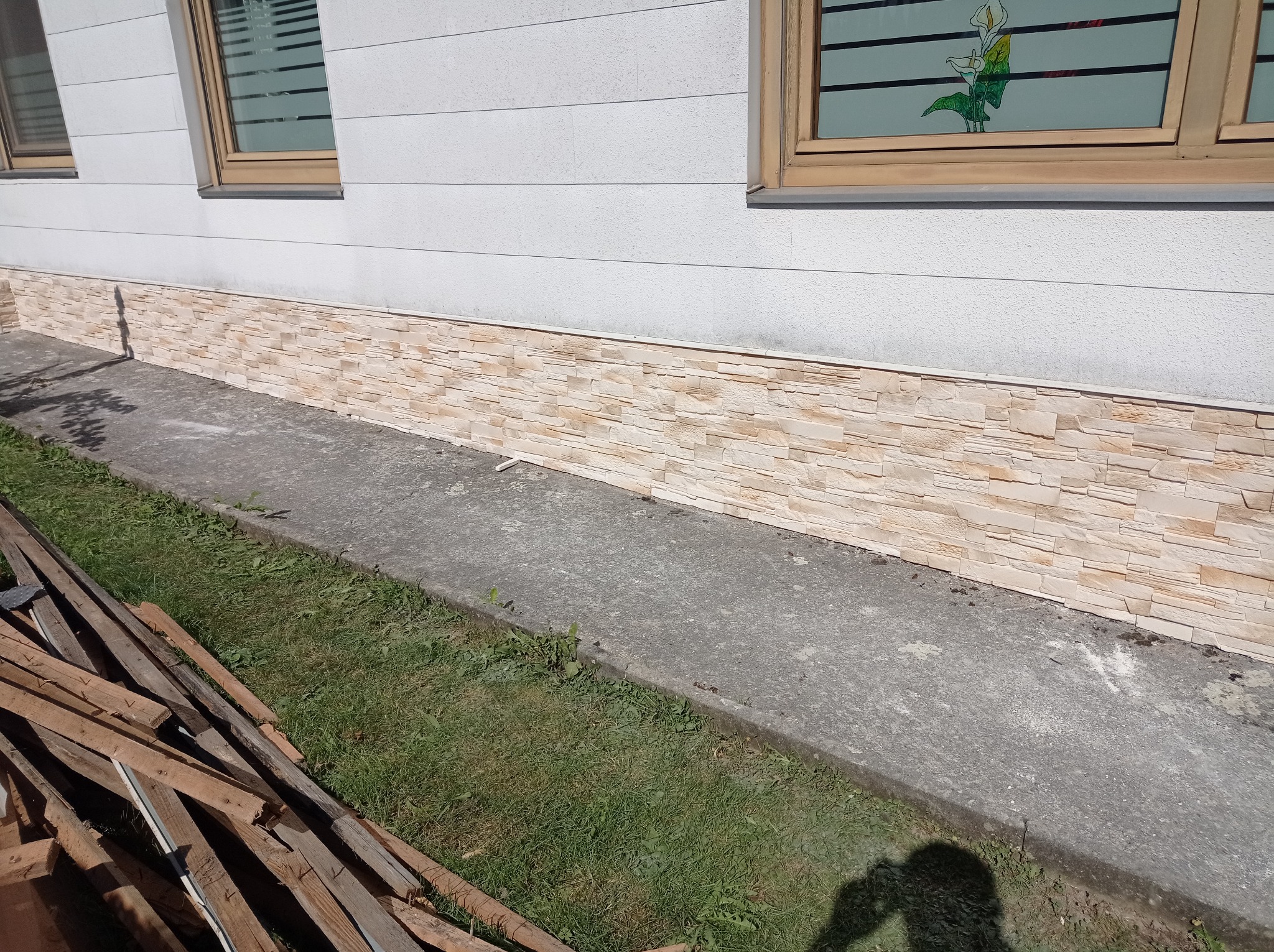 Wykończona elewacja domu z dekoracyjnym panelem imitującym kamień, widoczne okna z drewnianymi ramami i fragment trawnika z drewnianymi elementami konstrukcyjnymi.