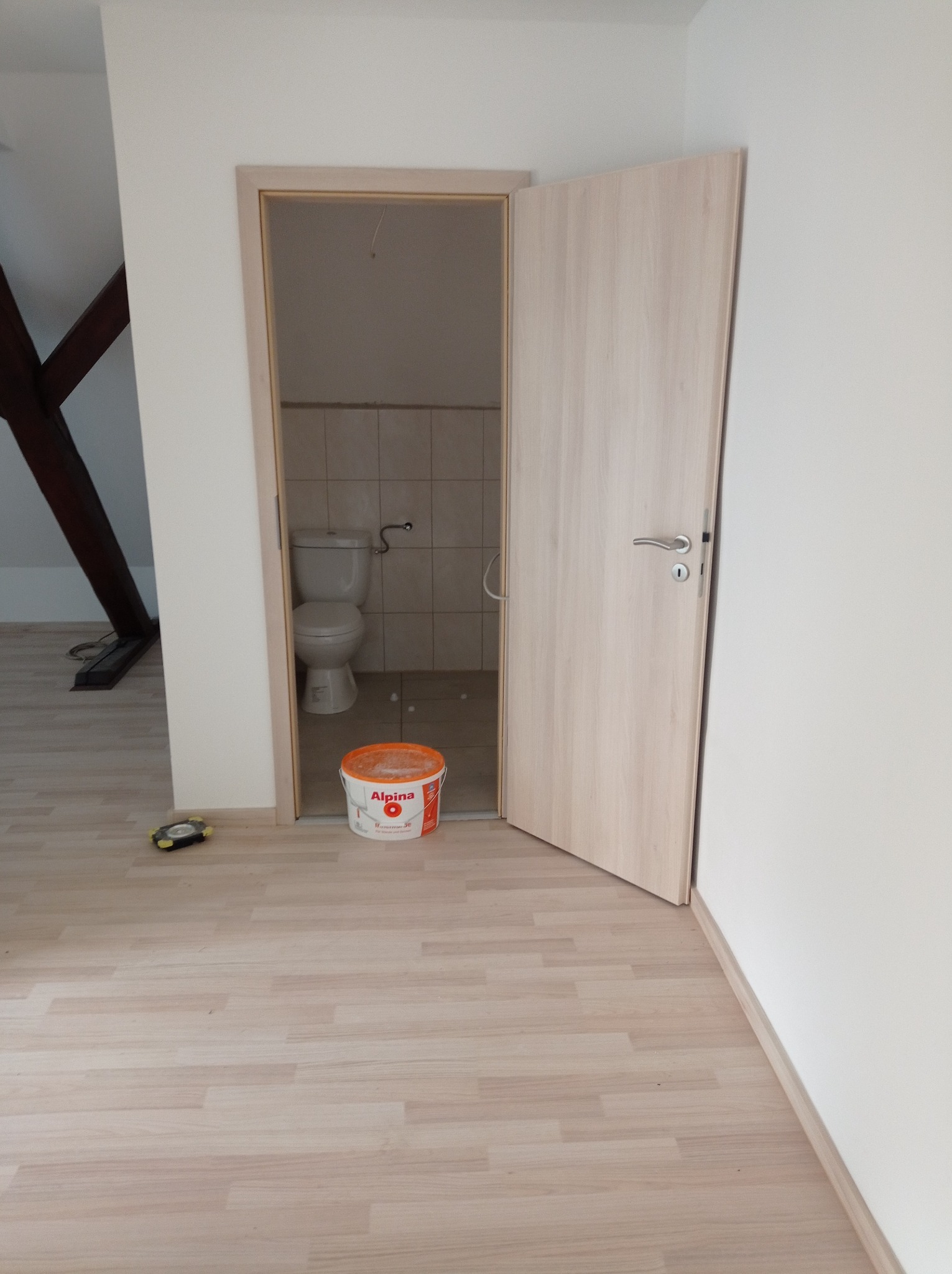 Otwarta, jasna drewniana wewnętrzna ościeżnica z drzwiami, przez którą widać wykafelkowaną toaletę z muszlą klozetową; na podłodze stoi wiadro z farbą marki Alpina oraz mała lampka inspekcyjna.