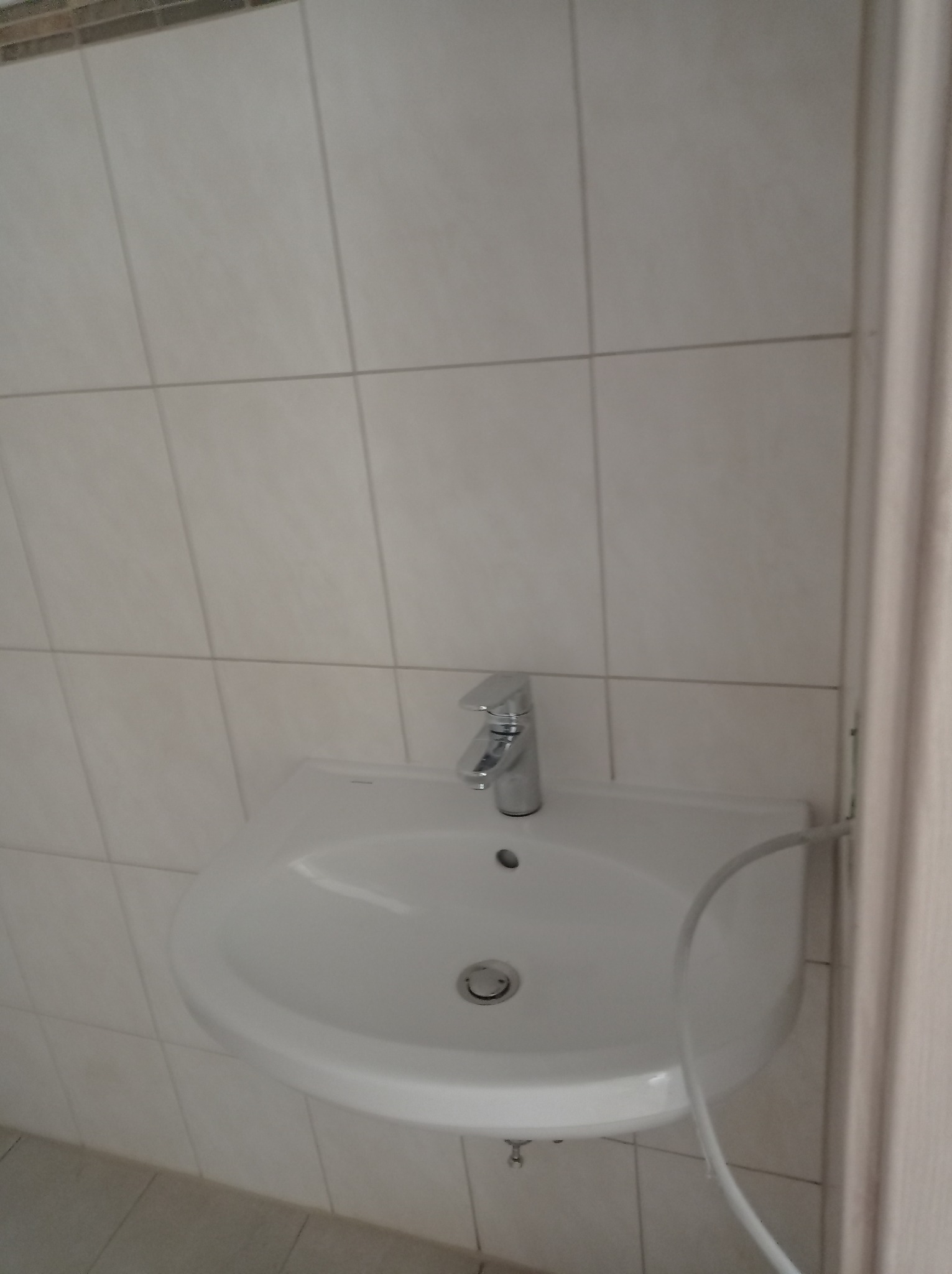 Biała umywalka z chromowaną baterią zamontowana na ścianie wyłożonej białymi, kwadratowymi płytkami ceramicznymi. Widoczny biały przewód doprowadzający wodę. Ujęcie z bliska, z widocznym fragmentem...