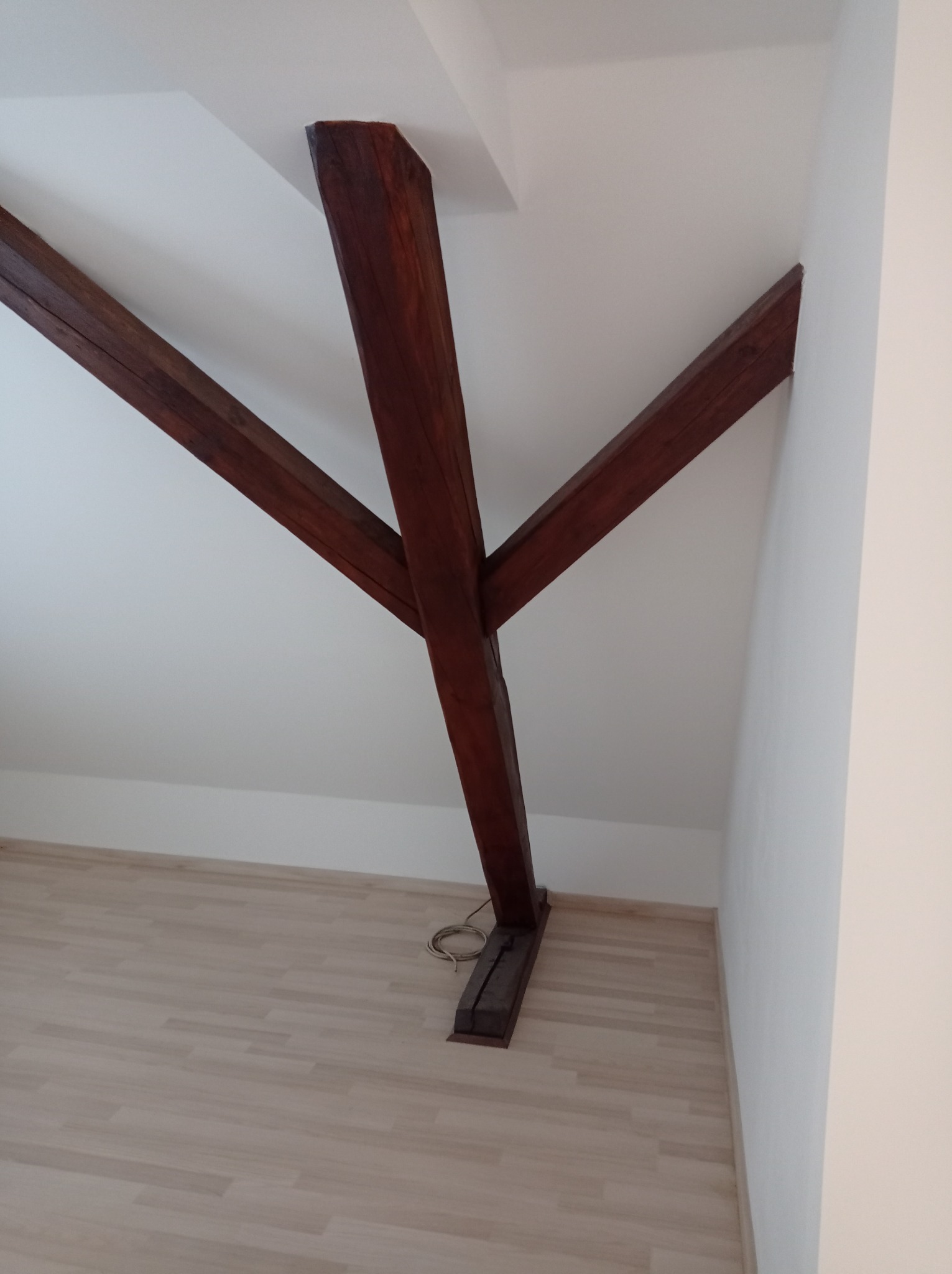 Wnętrze poddasza z drewnianą konstrukcją nośną w kształcie litery Y, bielone ściany i jasna podłoga, widoczne elementy instalacji elektrycznej przy podstawie słupa.