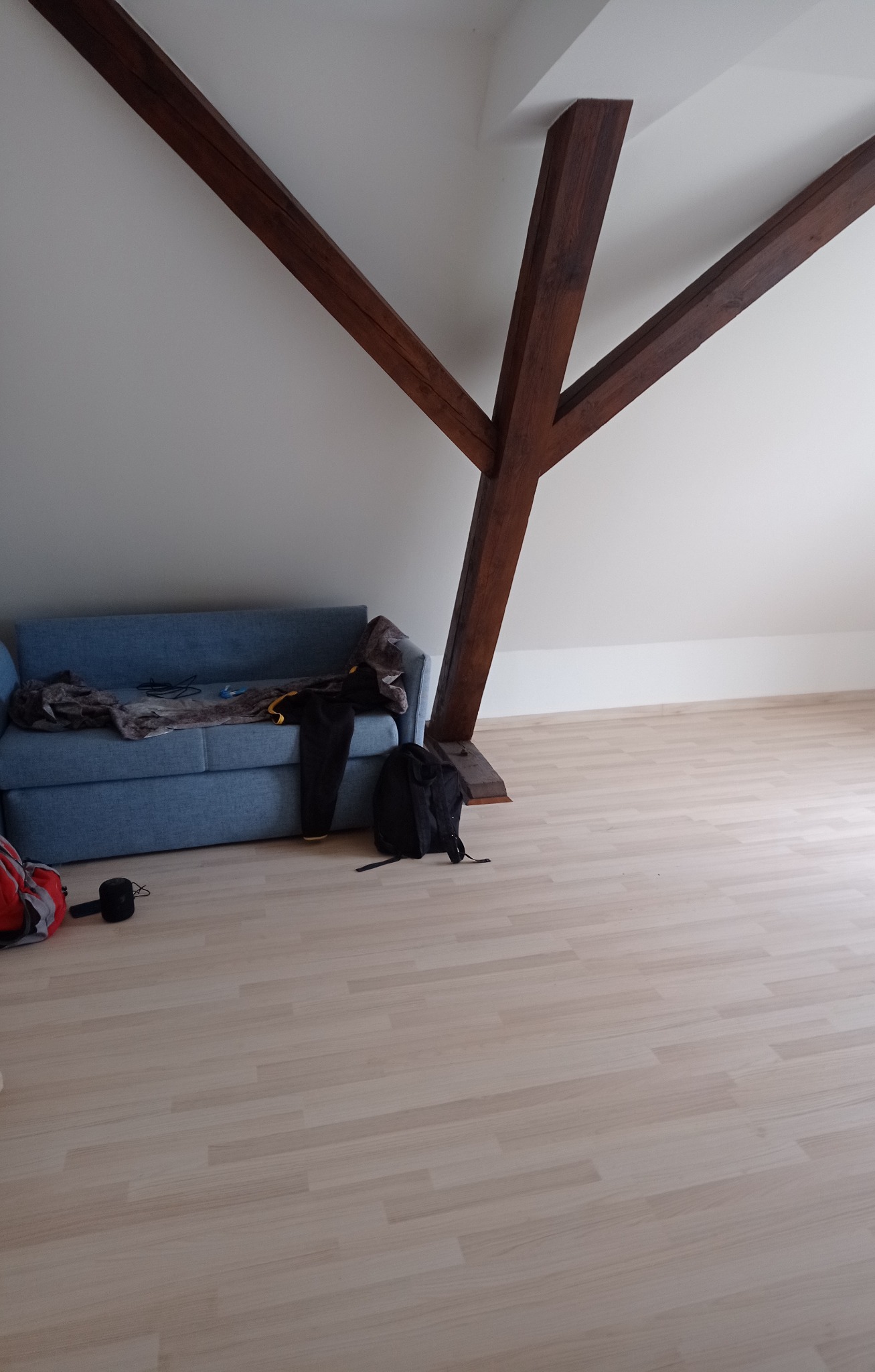 Poddasze z drewnianą konstrukcją nośną, niebieską rozkładaną sofą z rzeczami i plecakiem, jasną podłogą panelową i białymi ścianami.