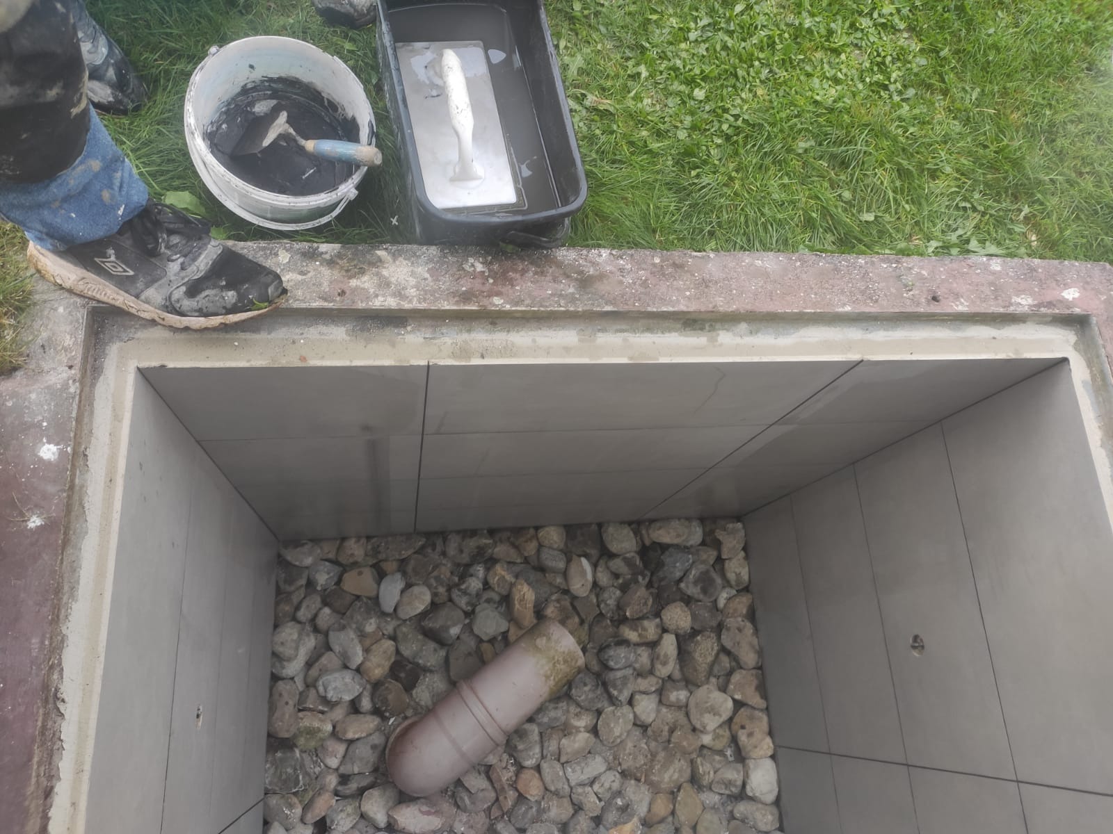 Wyłożony płytkami dół studzienki kanalizacyjnej z kamiennym dnem, widoczny kubeł z zaprawą, szpachelka i buty robocze na krawędzi.