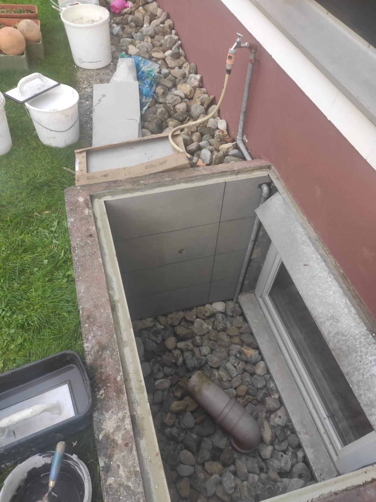 Wykończone okno piwniczne z szarą okładziną z płytek, drenażem z kamieni i rurą odpływową, widoczne narzędzia i materiały budowlane na zewnątrz.