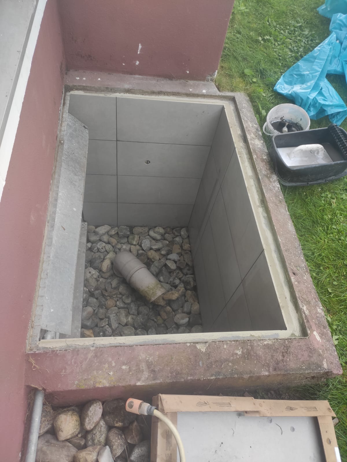 Otwarty właz do piwnicy wyłożony płytkami z kamienistym dnem i fragmentem rury odpływowej, widoczne narzędzia i materiały budowlane obok.