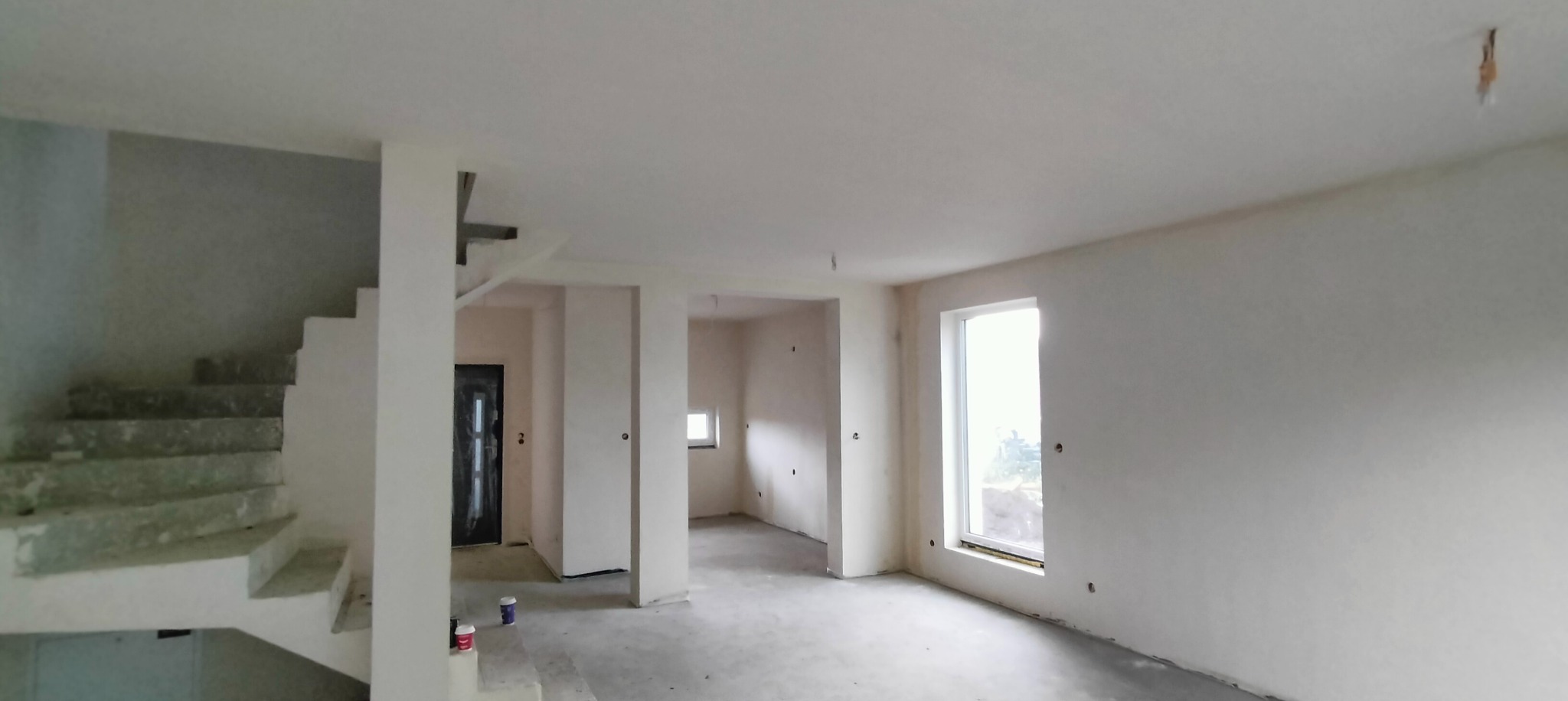 Surowe wnętrze domu w Szczecinie przed wykończeniem: surowe schody, świeżo zagruntowane ściany, widoczne puszki z farbą, duże okno z widokiem na zewnątrz.