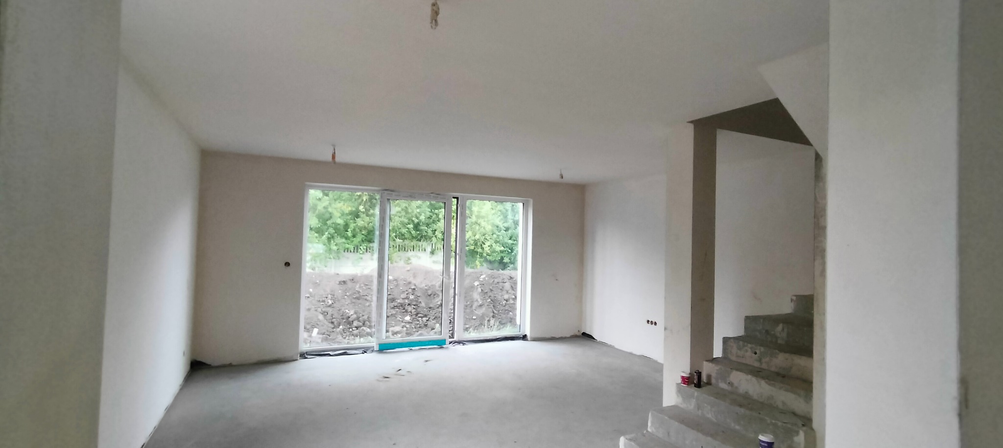 Surowe wnętrze z dużym oknem tarasowym, betonowa podłoga, otynkowane ściany, widoczne przygotowania do dalszych prac wykończeniowych przy schodach.