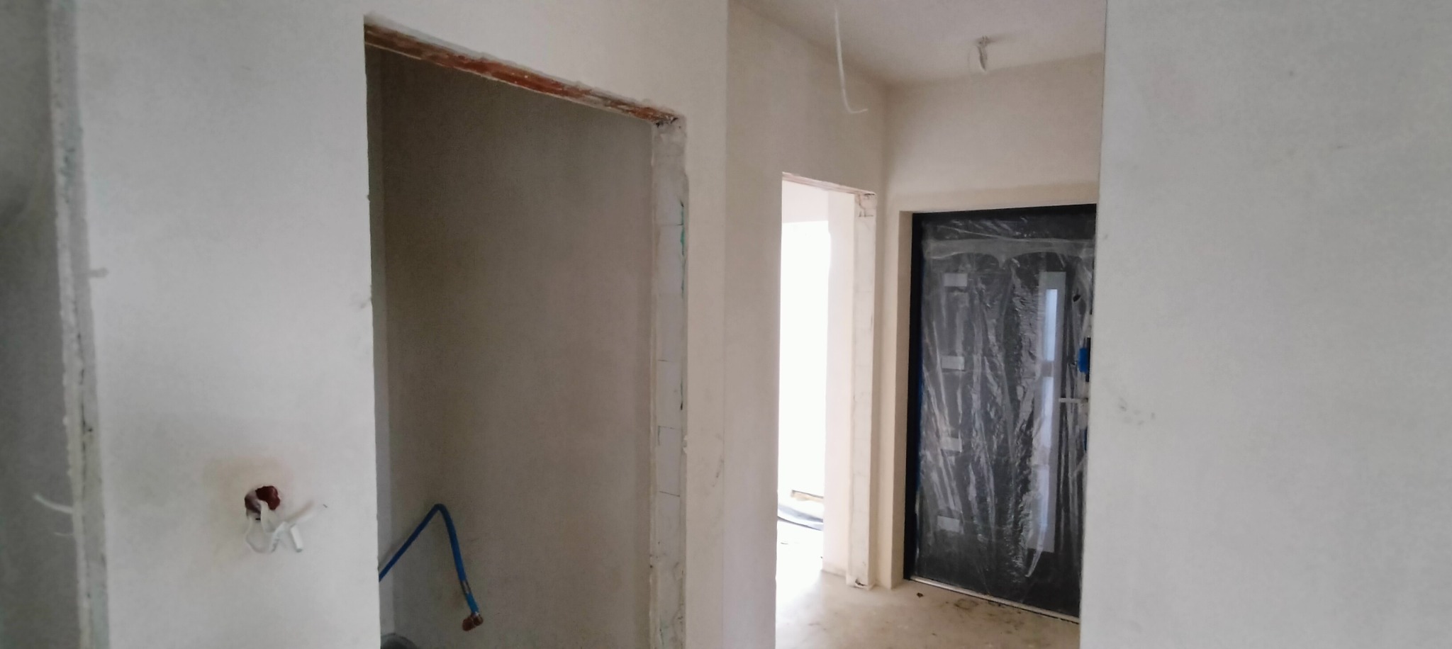 Wnętrze w trakcie prac wykończeniowych: surowe ściany, instalacja hydrauliczna wystająca ze ściany, drzwi wejściowe zabezpieczone folią ochronną.