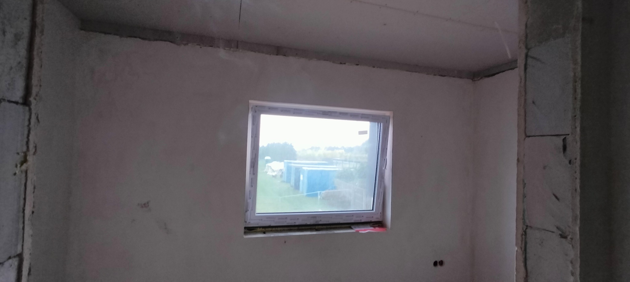 Surowy stan wnętrza z oknem, ściany po nałożeniu gładzi, widoczne ślady po łatach, sufit w stanie surowym, bloczki betonowe przy otworze drzwiowym.