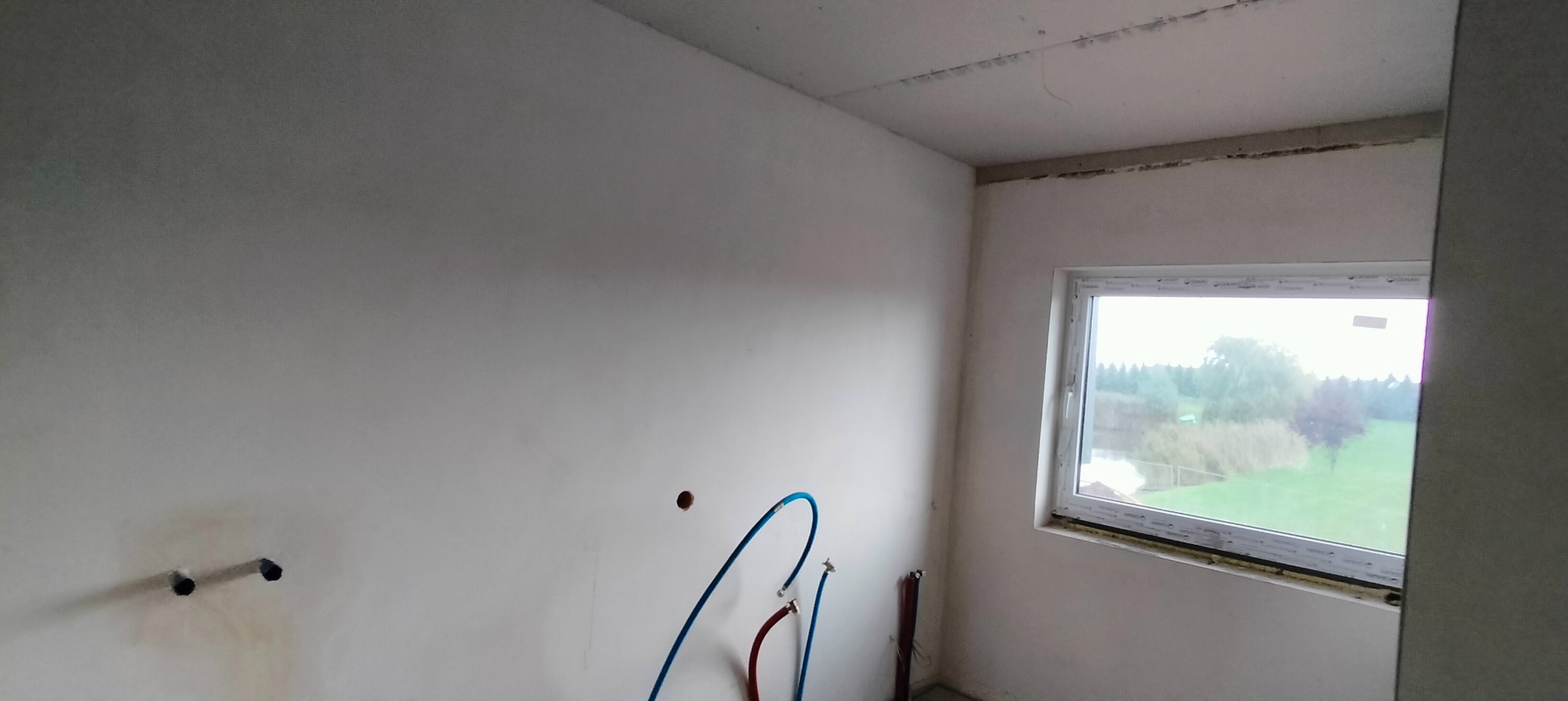 Surowa ściana z instalacją hydrauliczną w pokoju z oknem, widoczne niezagruntowane ściany i sufit przed wykończeniem.