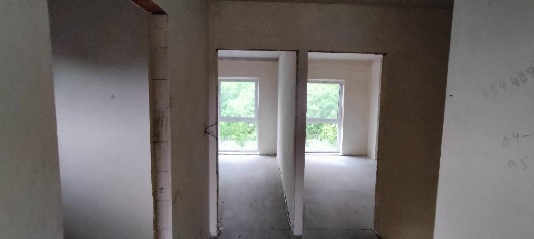 Surowe wnętrze z dwoma oknami i ścianami w trakcie wykończenia; widoczne ślady po instalacjach i notatki na ścianie.