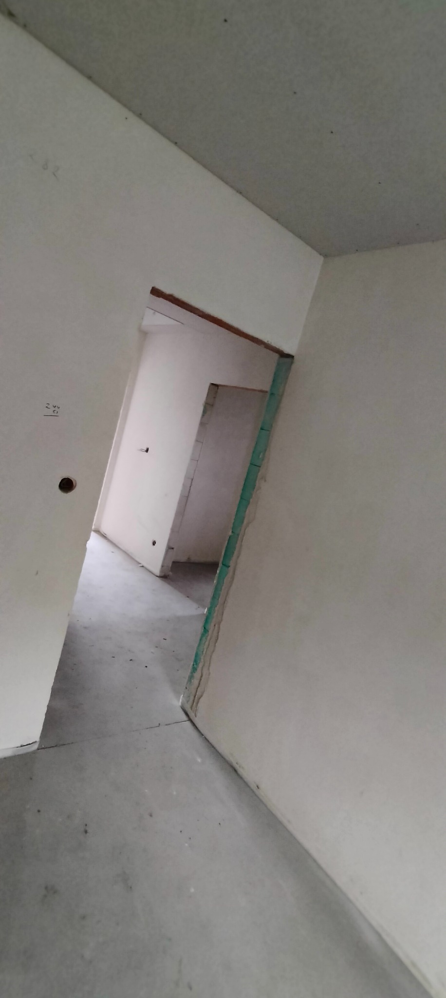 Wnętrze w trakcie prac wykończeniowych: surowe ściany, betonowa podłoga, widoczne profile i płyty gipsowo-kartonowe przy otworze drzwiowym. Na ścianie widoczne ślady po ołówku i otwór po puszce...