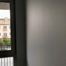 gotowesciany.pl - Świeżo pomalowana, gładka, szara ściana wewnętrzna z widokiem na balkon i budynek mieszkalny przez okno.