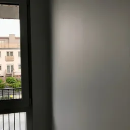 Świeżo pomalowana, gładka, szara ściana wewnętrzna z widokiem na balkon i budynek mieszkalny przez okno.