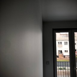 gotowesciany.pl - Świeżo pomalowana, gładka ściana w odcieniach szarości, widok przez balkon na budynek mieszkalny z drewnianymi schodami i zielonym ogrodem.