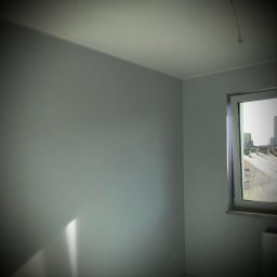 gotowesciany.pl - Pomalowane na szaro wnętrze pokoju z oknem, widok na zabudowę miejską, listwy przypodłogowe i narożniki wykończone, widoczny fragment grzejnika.