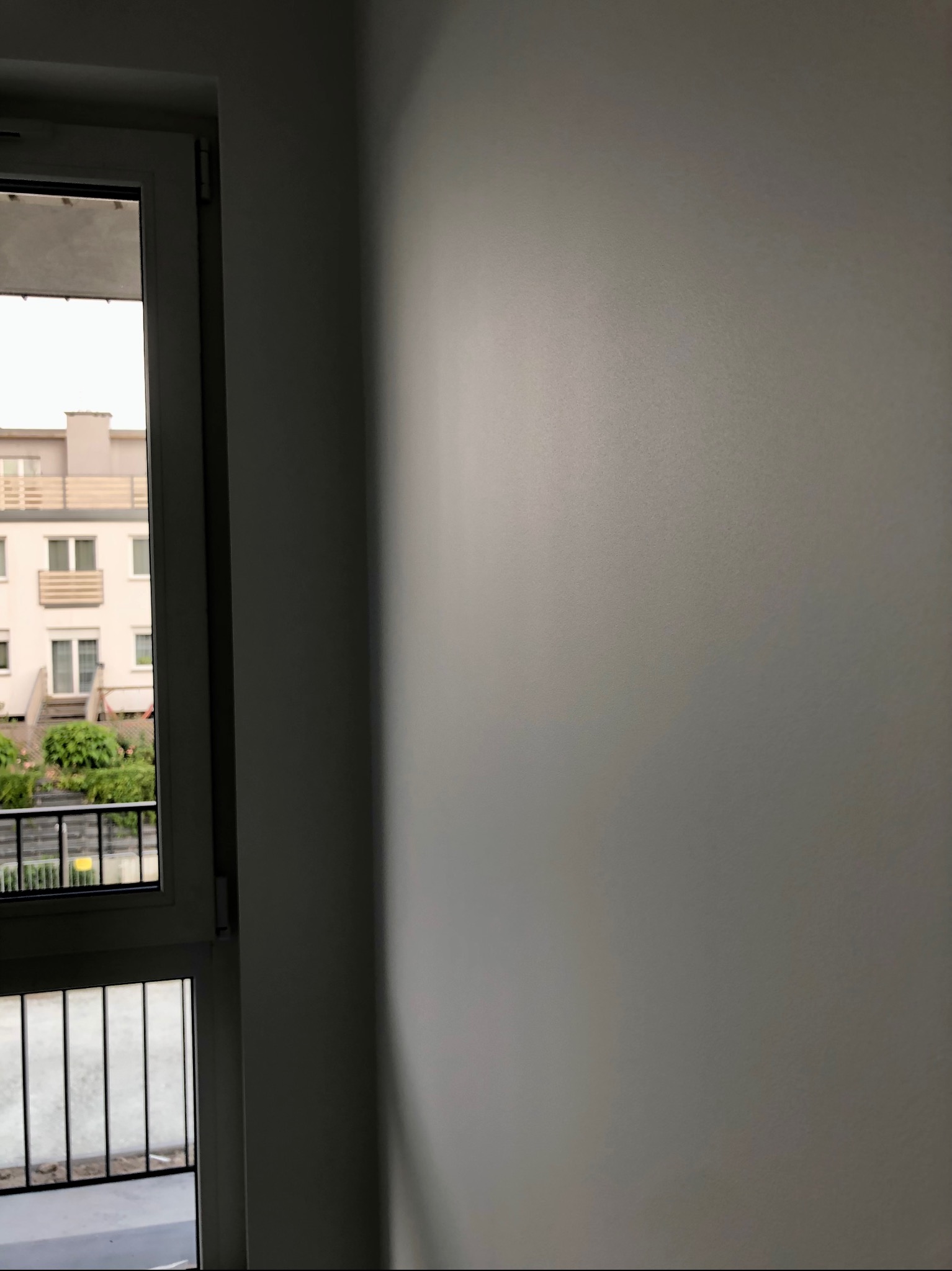 Świeżo pomalowana, gładka, szara ściana wewnętrzna z widokiem na balkon i budynek mieszkalny przez okno.