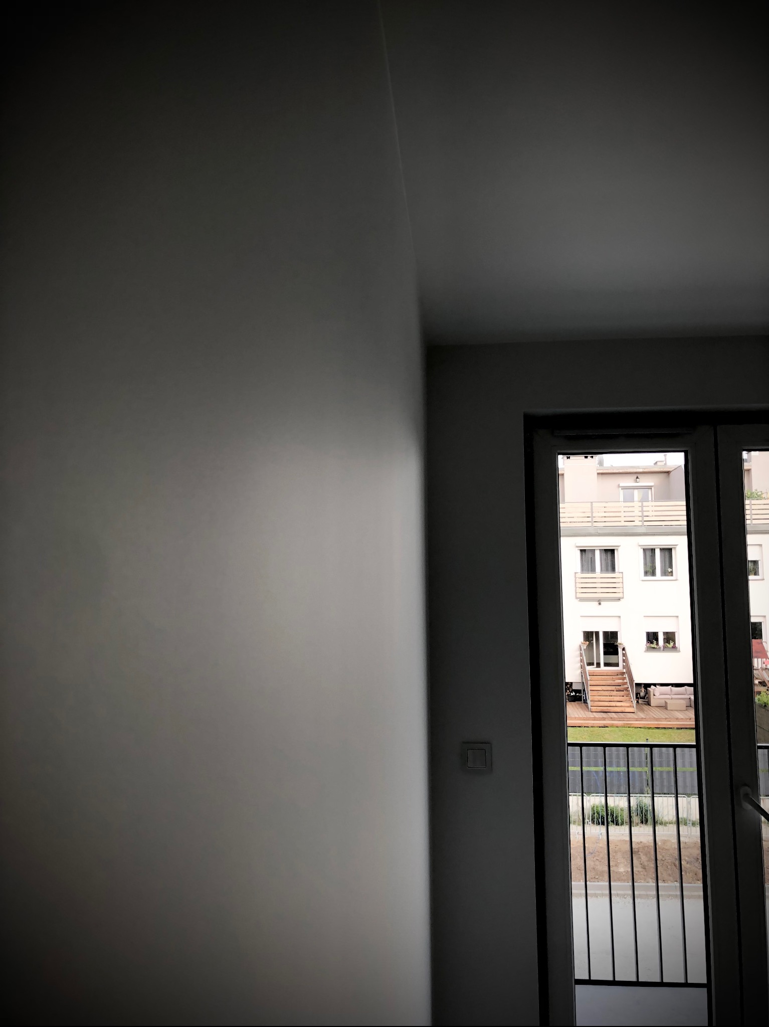 Świeżo pomalowana, gładka ściana w odcieniach szarości, widok przez balkon na budynek mieszkalny z drewnianymi schodami i zielonym ogrodem.