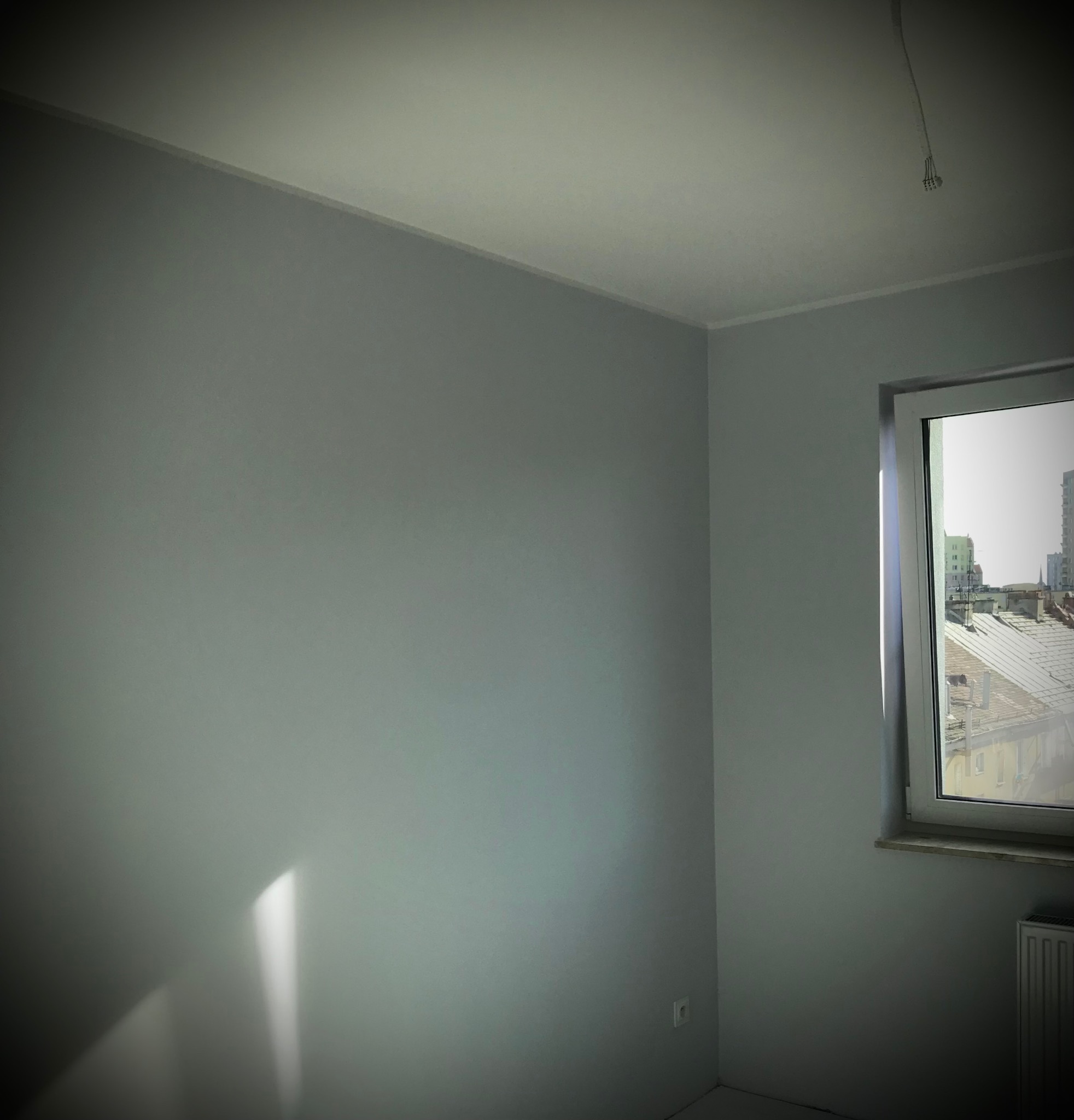 Pomalowane na szaro wnętrze pokoju z oknem, widok na zabudowę miejską, listwy przypodłogowe i narożniki wykończone, widoczny fragment grzejnika.