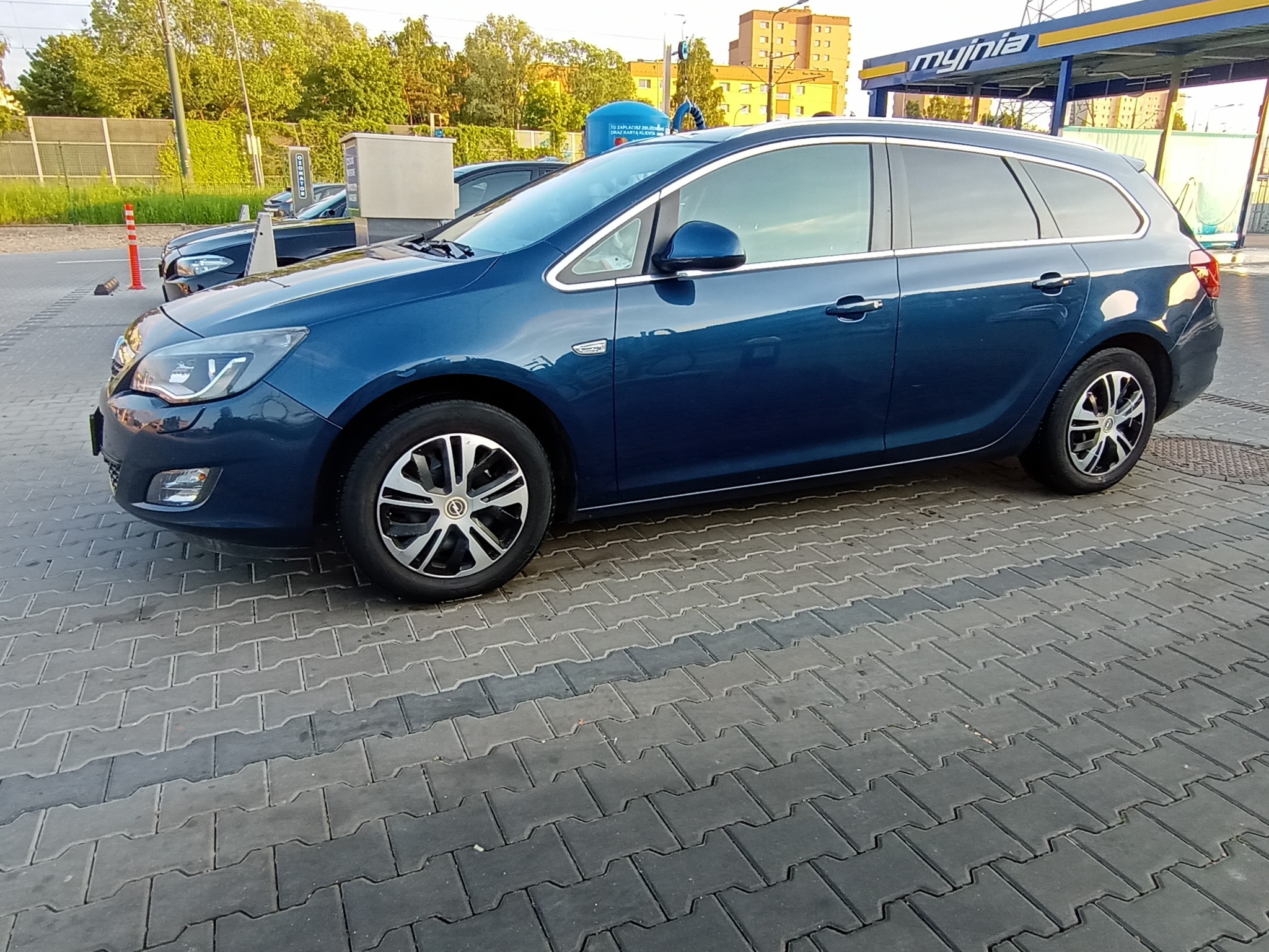 Niebieski samochód kombi marki Opel zaparkowany na kostce brukowej przed myjnią samochodową z żółto-niebieskim szyldem.