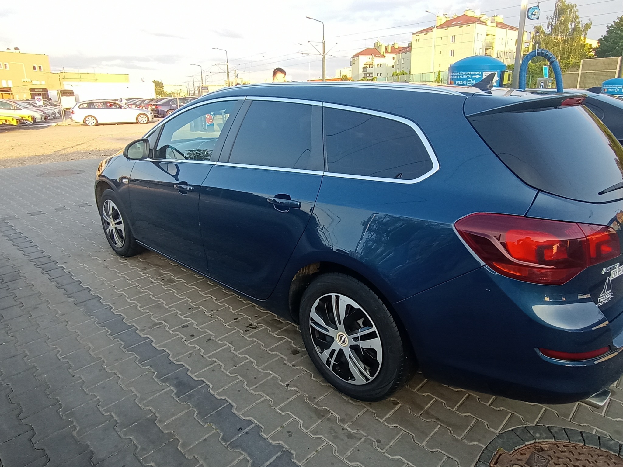 Niebieskie kombi Opel Astra zaparkowane na chodniku z kostki brukowej, widok z tyłu z boku, w tle budynki i myjnia samochodowa.