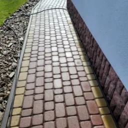 Mokra kostka brukowa w odcieniach brązu i beżu, ułożona wzdłuż ściany budynku z cokołem z kamienia dekoracyjnego, z widocznym przewodem zasilającym.