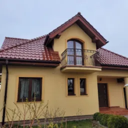 Żółta elewacja domu jednorodzinnego z brązowymi oknami i dachem z dachówki ceramicznej, widok z poziomu trawnika.