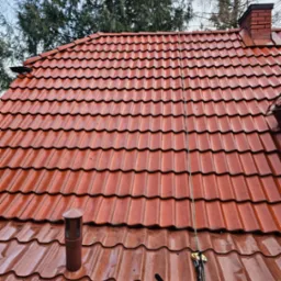 Czerwona dachówka ceramiczna, lekko wilgotna, z widocznym kominem i liną zabezpieczającą, sugerującą prace na wysokości.