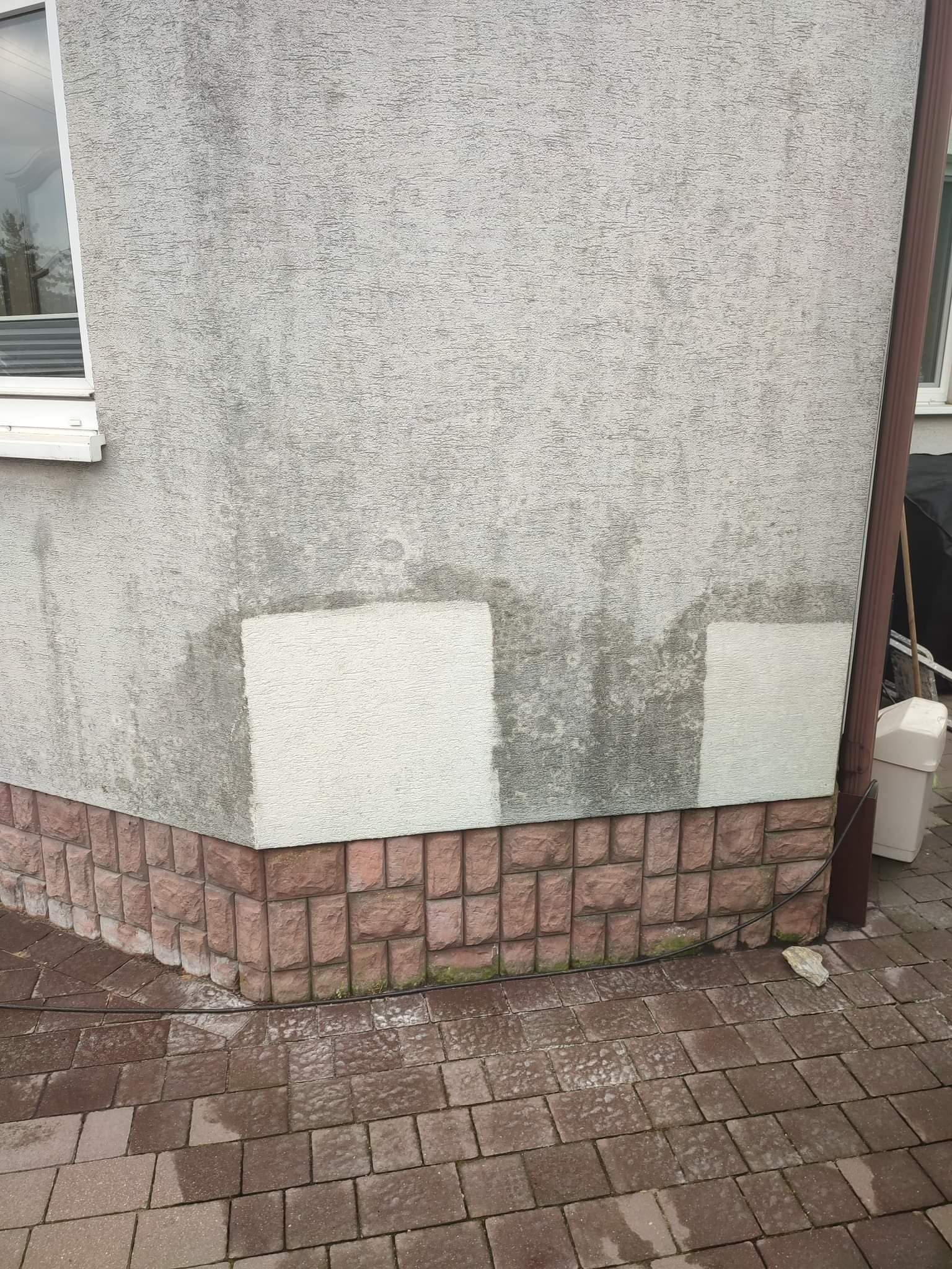 Fragment narożnika budynku z szarą, zabrudzoną elewacją z widocznymi śladami po czyszczeniu, z ceglaną podmurówką i mokrą kostką brukową.