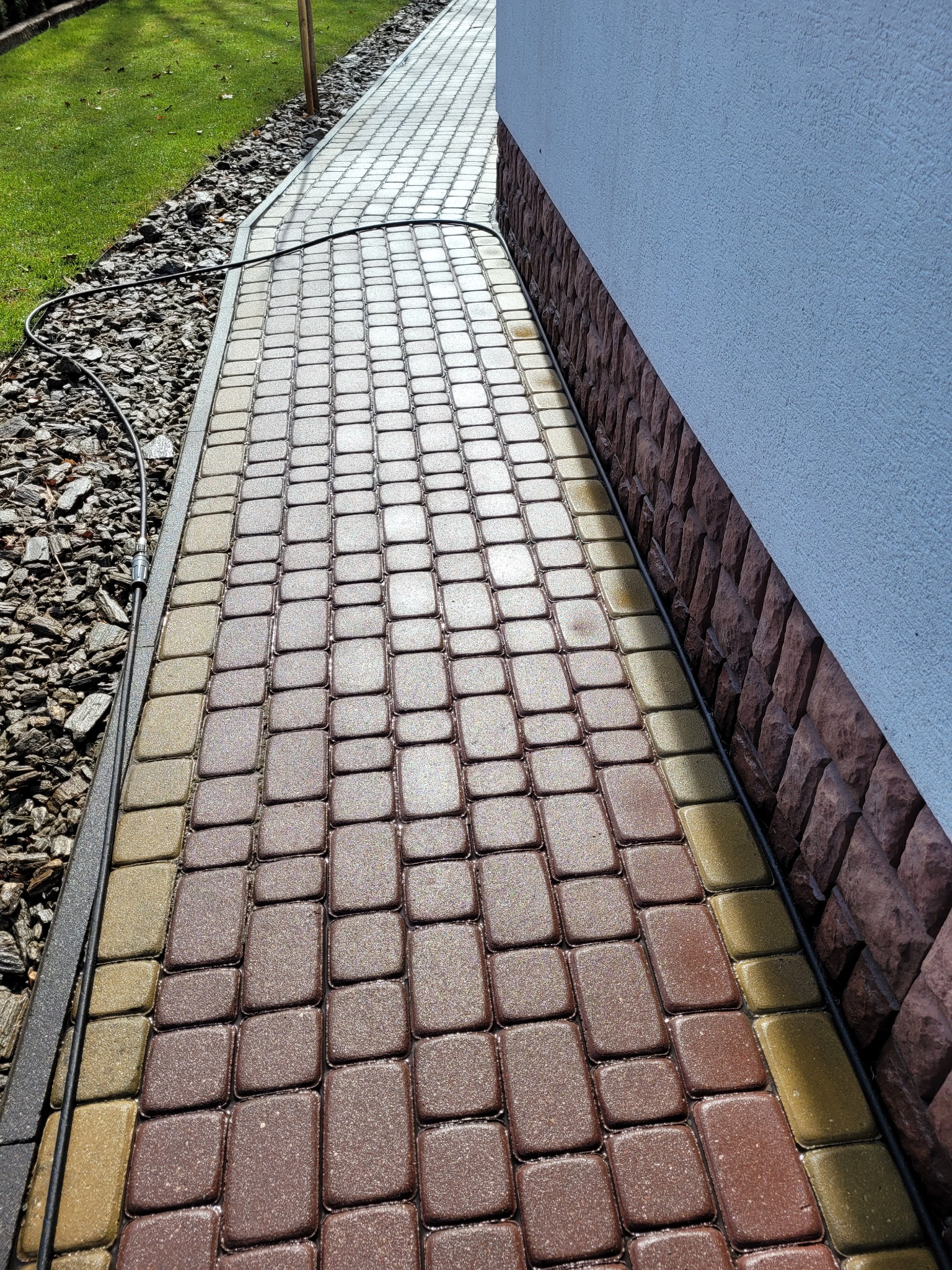 Mokra kostka brukowa w odcieniach brązu i beżu, ułożona wzdłuż ściany budynku z cokołem z kamienia dekoracyjnego, z widocznym przewodem zasilającym.