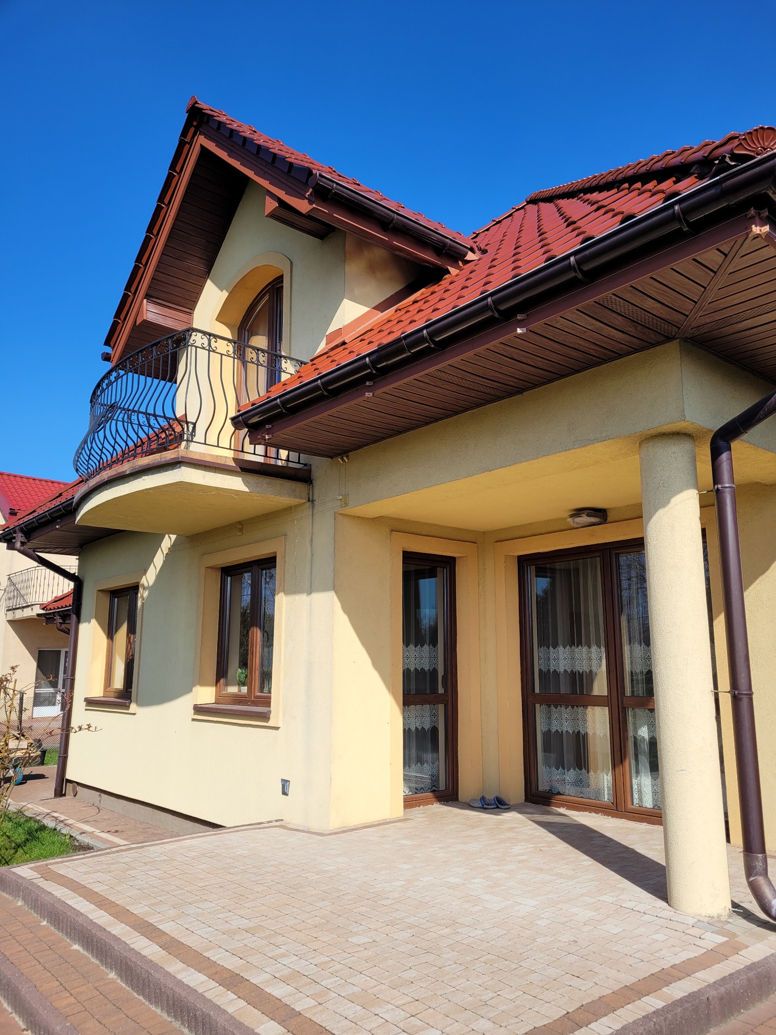 Dwupiętrowy dom z żółtą elewacją, czerwonym dachem i balkonem z kutą balustradą, widziany w słoneczny dzień.