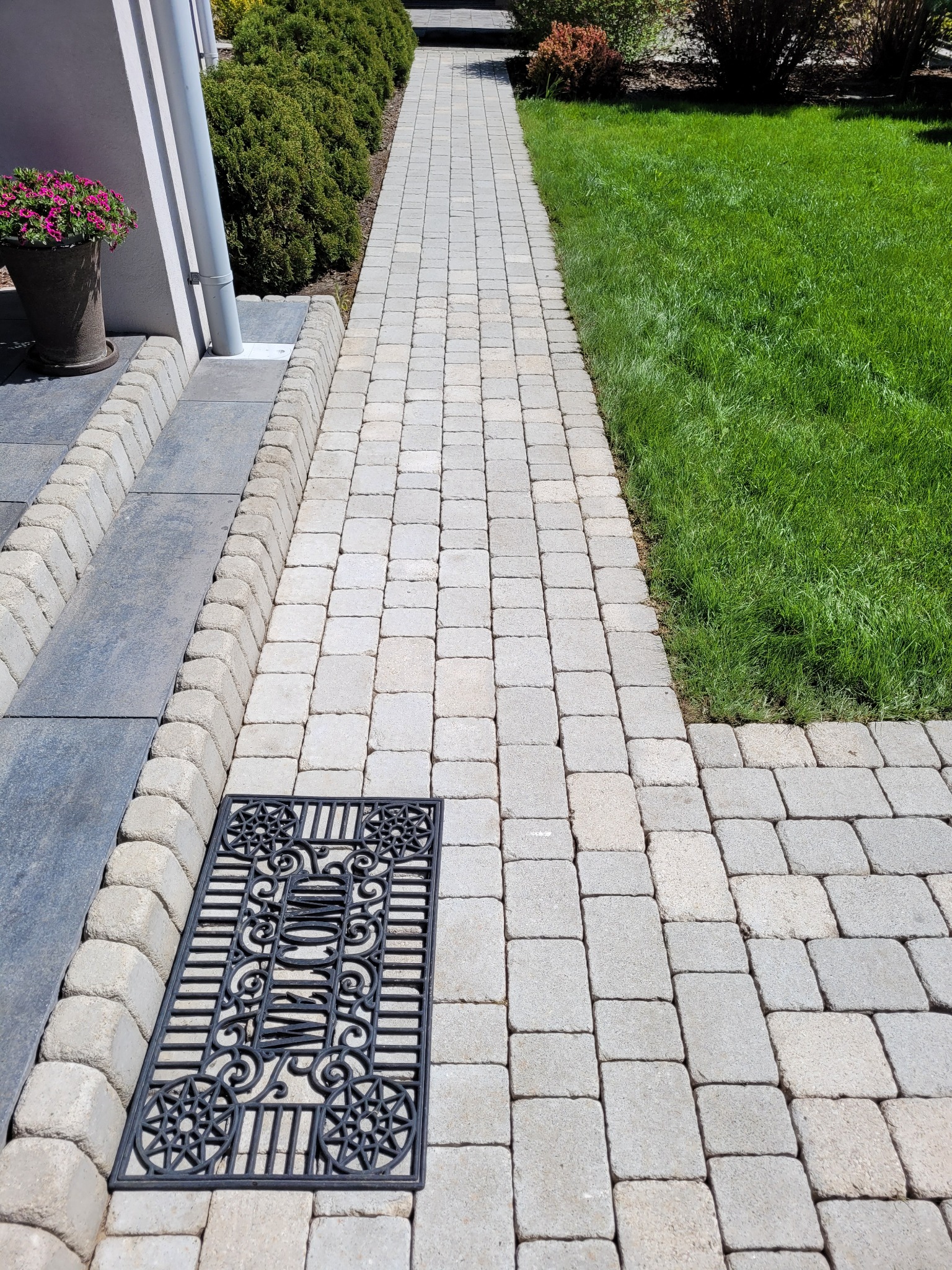 Ułożona kostka brukowa tworząca ścieżkę z ozdobną wycieraczką przed wejściem do domu z zielonym trawnikiem po prawej stronie.