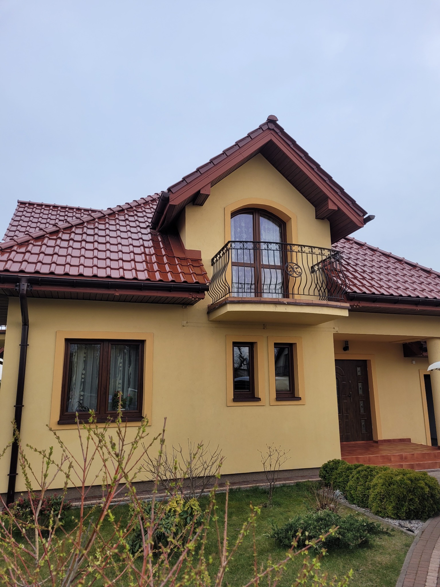 Żółta elewacja domu jednorodzinnego z brązowymi oknami i dachem z dachówki ceramicznej, widok z poziomu trawnika.