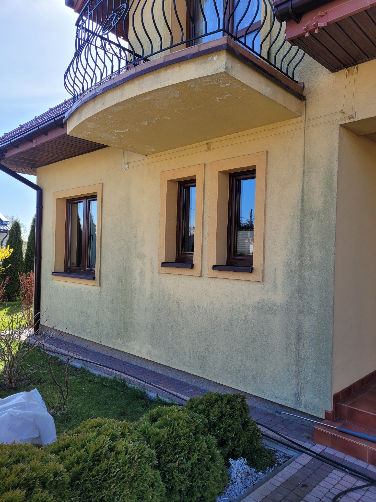 Żółta elewacja domu z trzema oknami o brązowych ramach, widoczne ślady zabrudzeń, balkon z czarną, ozdobną balustradą i fragment dachu z brązową dachówką.
