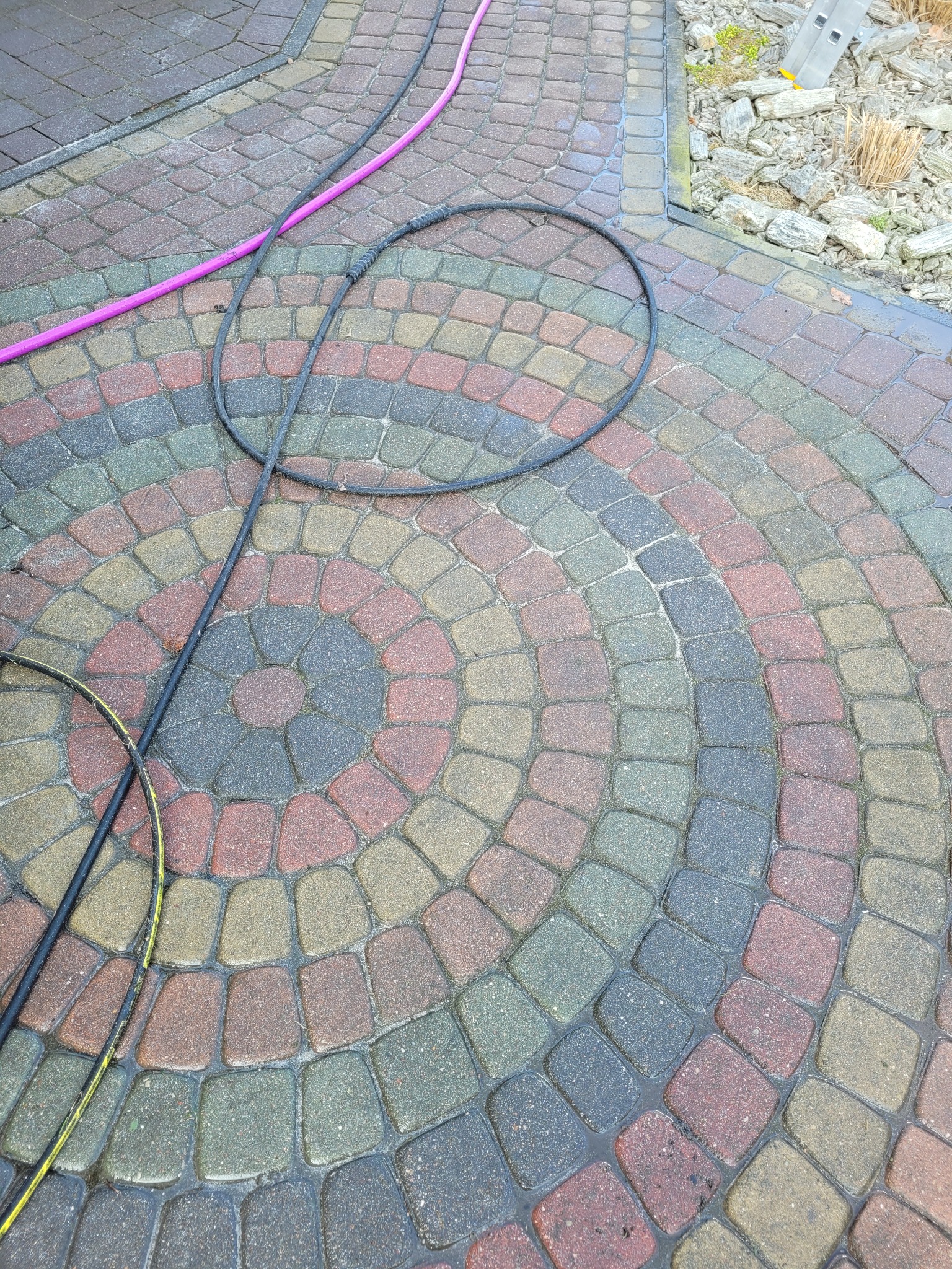 Mokra kostka brukowa ułożona w koncentryczny wzór, z widocznymi wężami ciśnieniowymi w kolorze różowym i czarnym, oraz fragment drabiny w tle, sugerujące prace porządkowe lub czyszczenie.