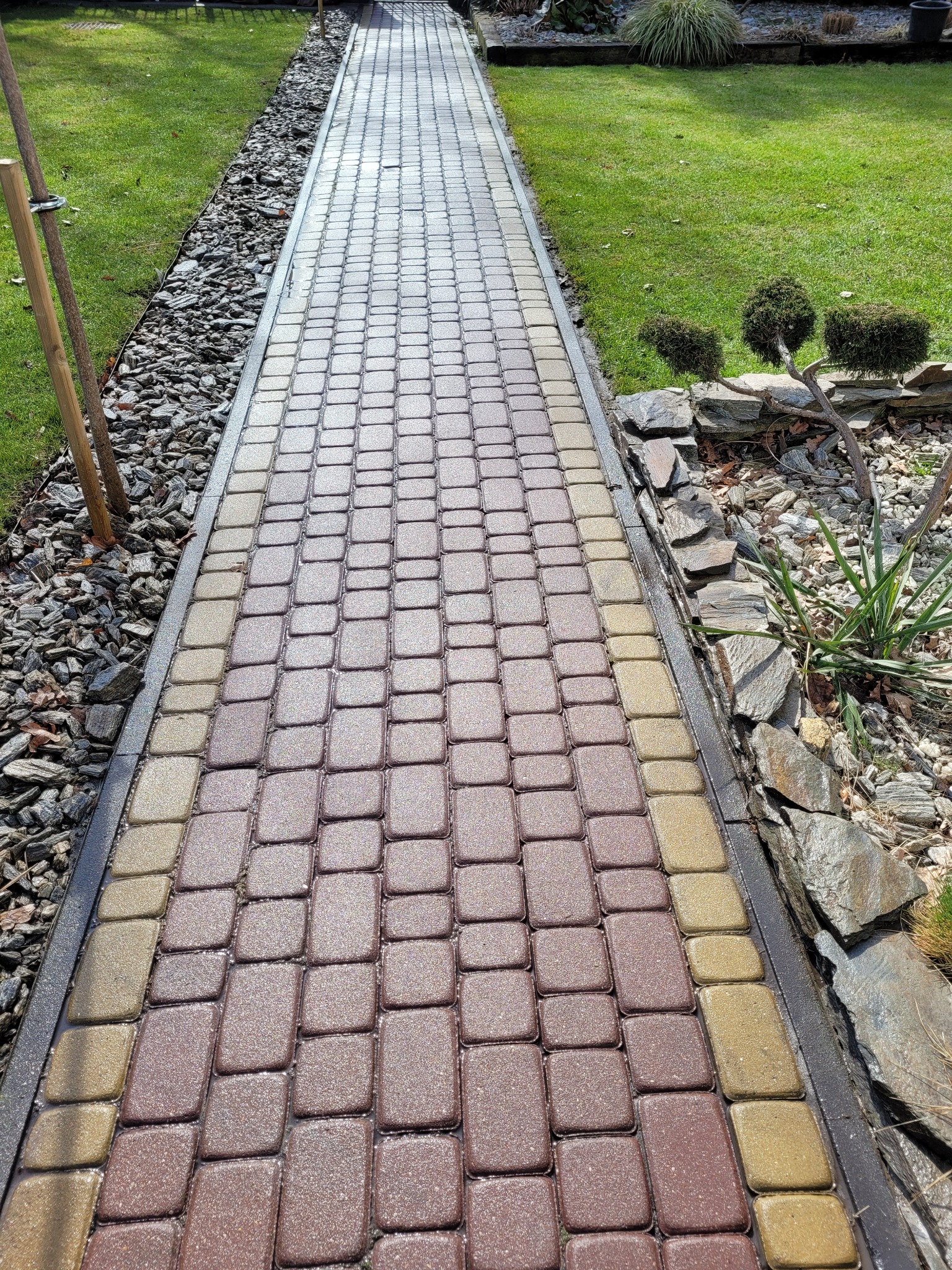 Długa ścieżka z kostki brukowej w odcieniach brązu i beżu, obrzeża wykończone kamieniami i ozdobnym żwirem, prowadząca przez zadbany trawnik z formowanymi krzewami.