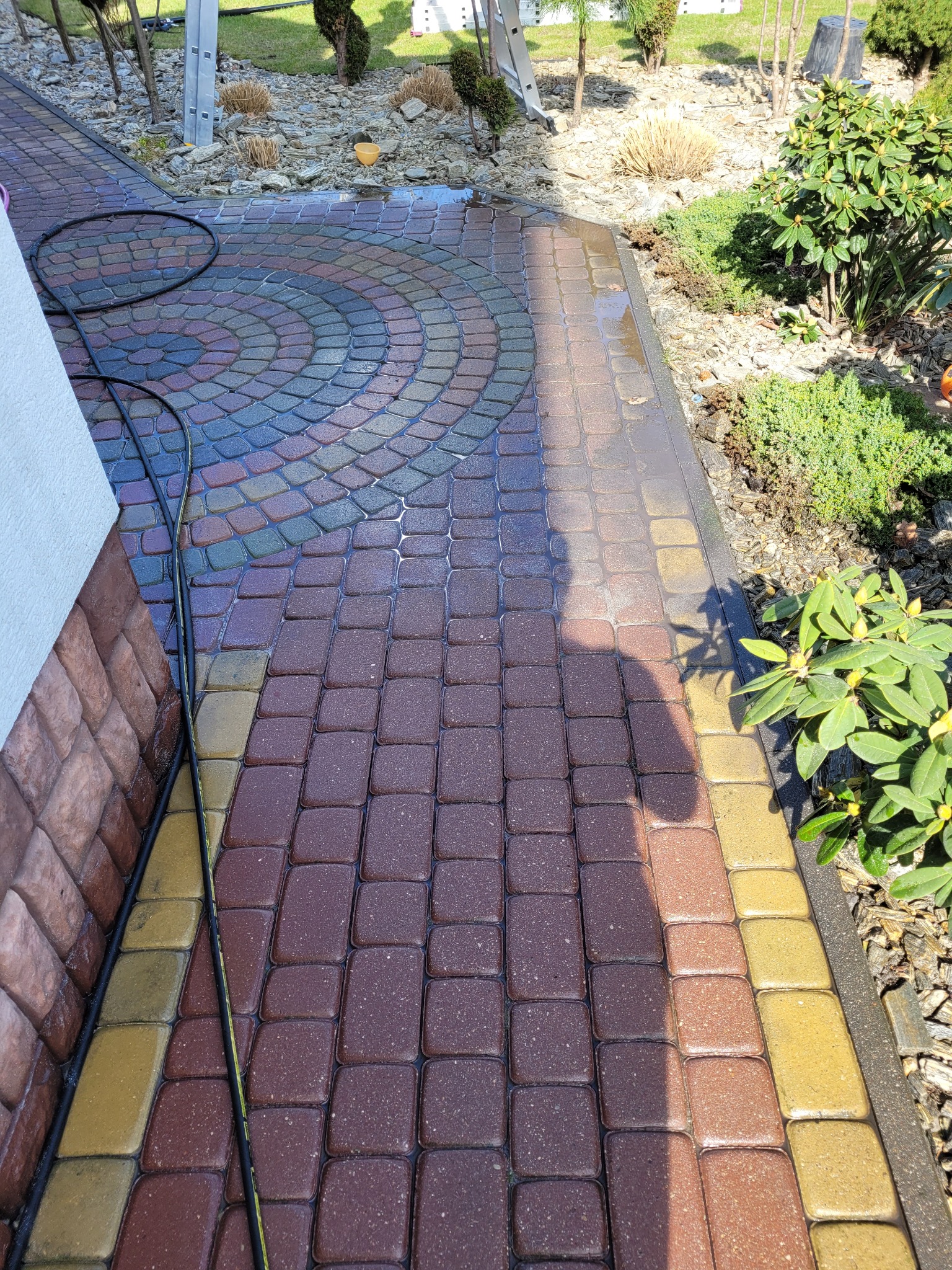Częściowo umyta, wielobarwna kostka brukowa z widocznym cieniem osoby, w tle roślinność i fragment elewacji budynku.