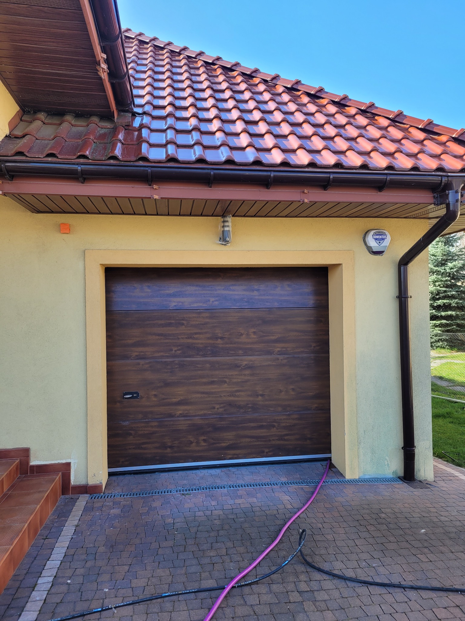 Fragment elewacji domu z brązową bramą garażową i częściowo umytym dachem z czerwonej dachówki, widoczny wąż ogrodowy na kostce brukowej.